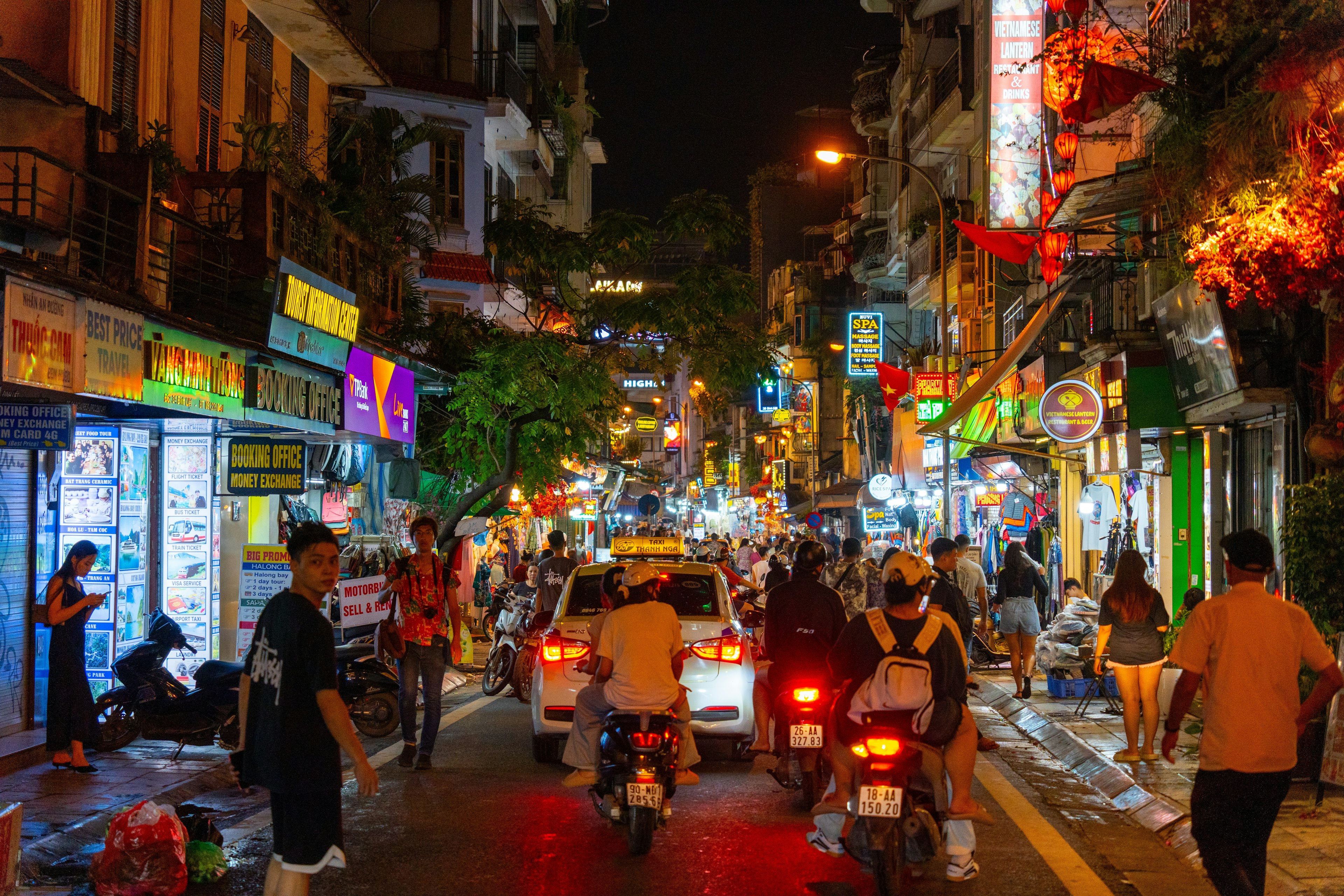 A narrow road full of cars and motorbikes at night. People walk along the sidewalks and behind the vehicles.