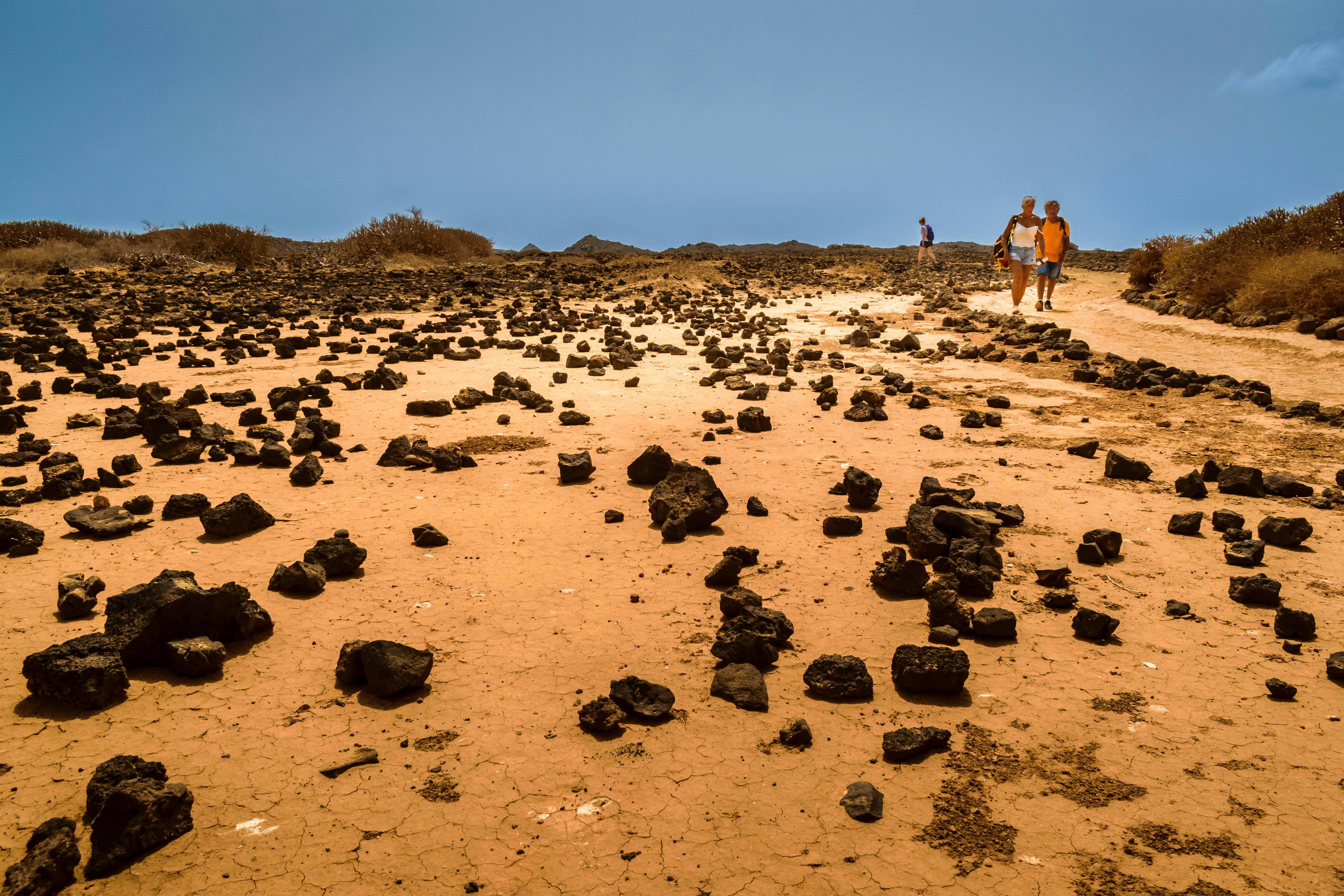 Rocks amid sand with two people walking nearby and dunes in the background