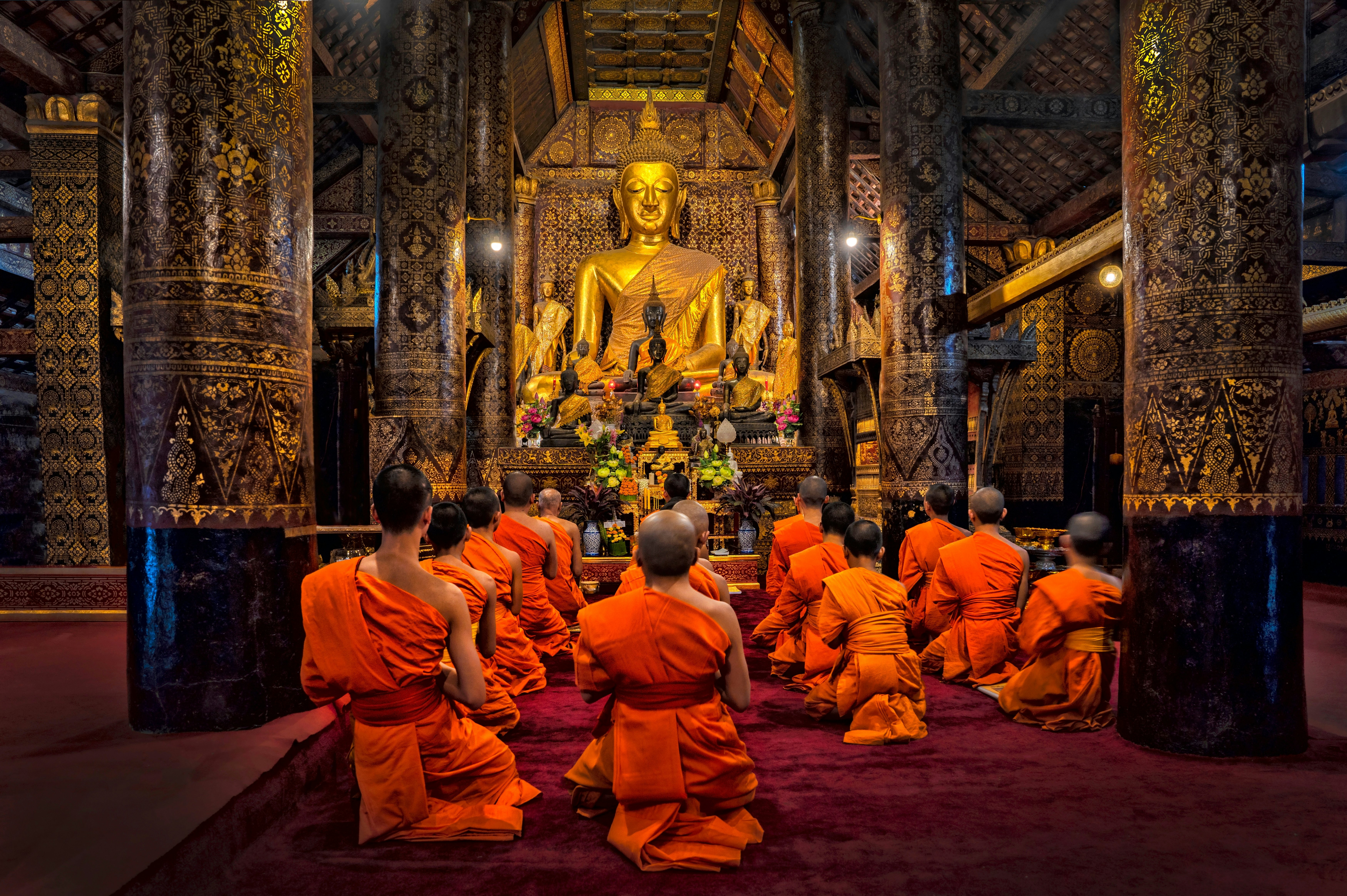 Orange-robed monks sit in prayer in a temple with a large golden Buddha statue.