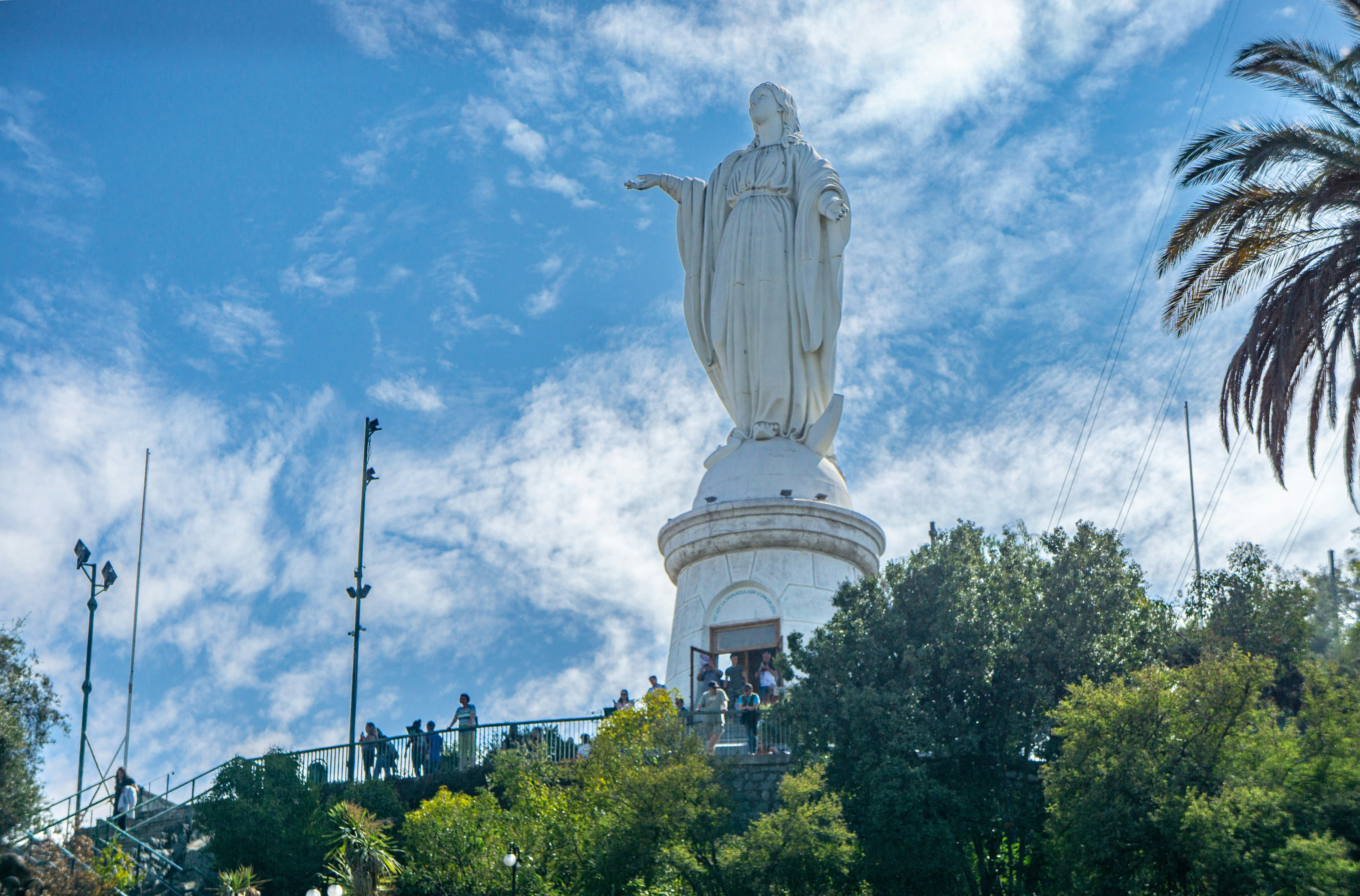 A huge statue of the Virgin Mary at the top of a hill in a city park, with people standing at an overlook in front.