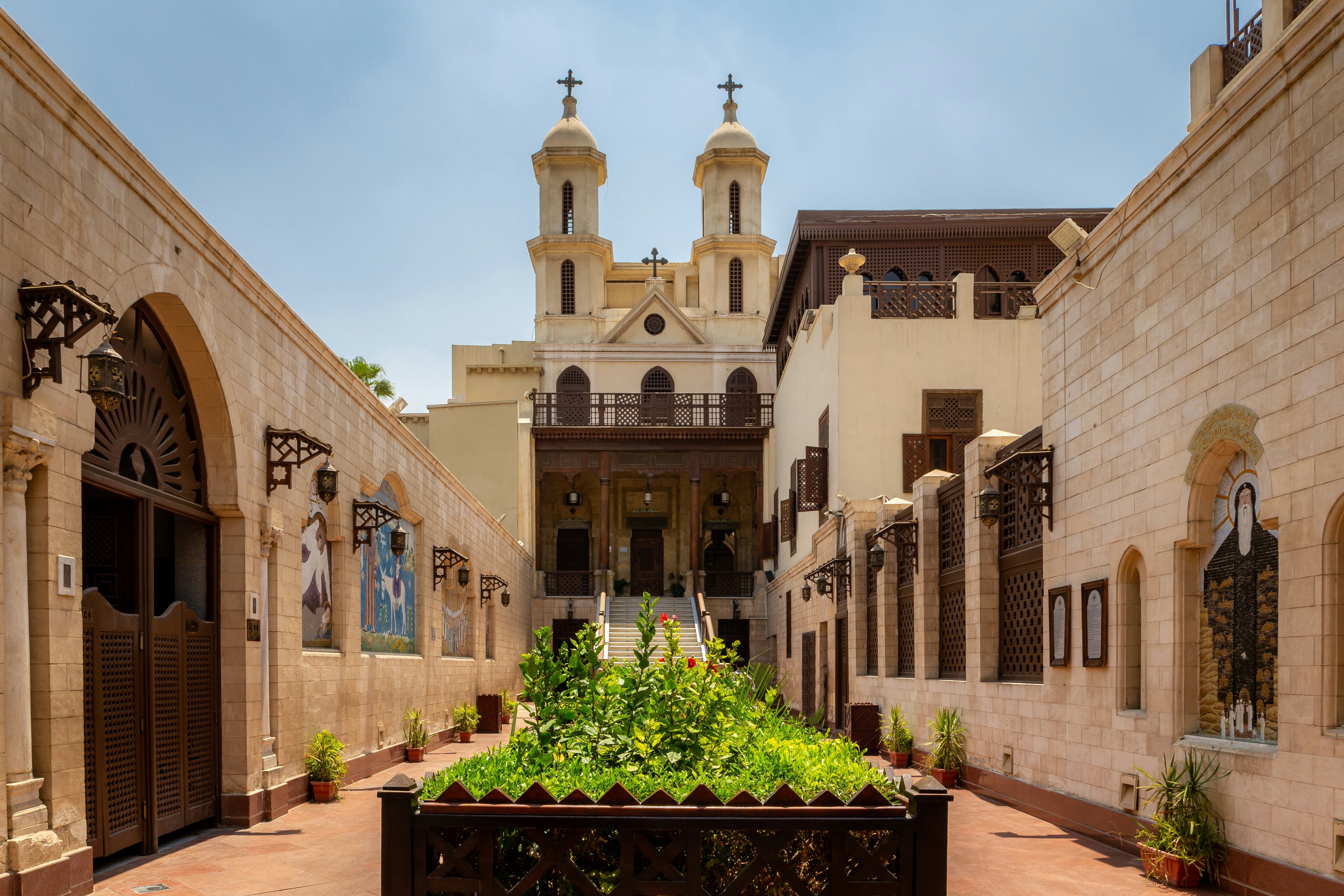 A white church with dark accents is reached by a staircase from a narrow alley with a garden bed in the center.