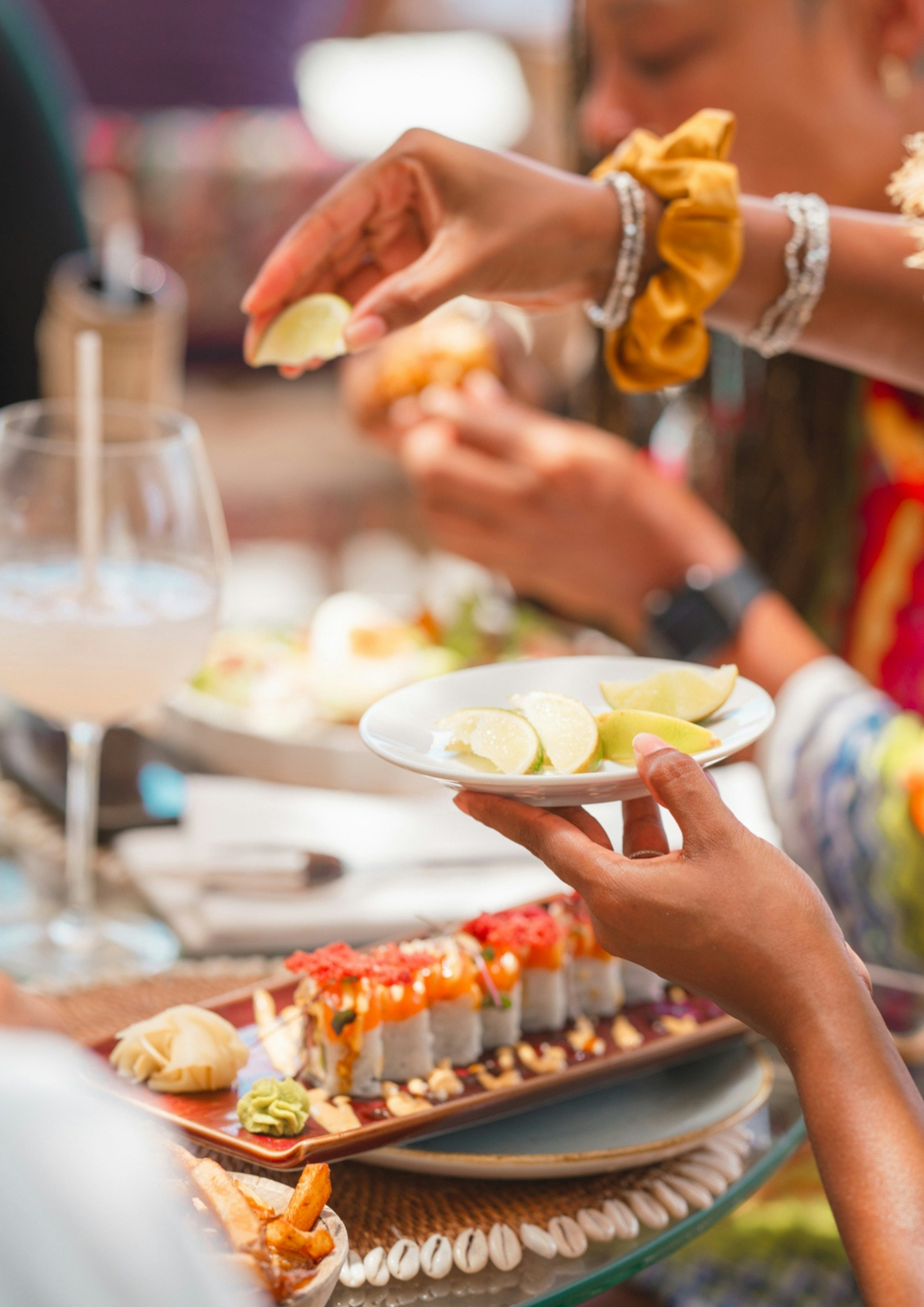 A person's left hand holding slices of limes over a platter of sushi while they squeeze lime juice into a cocktail glass with their right hand