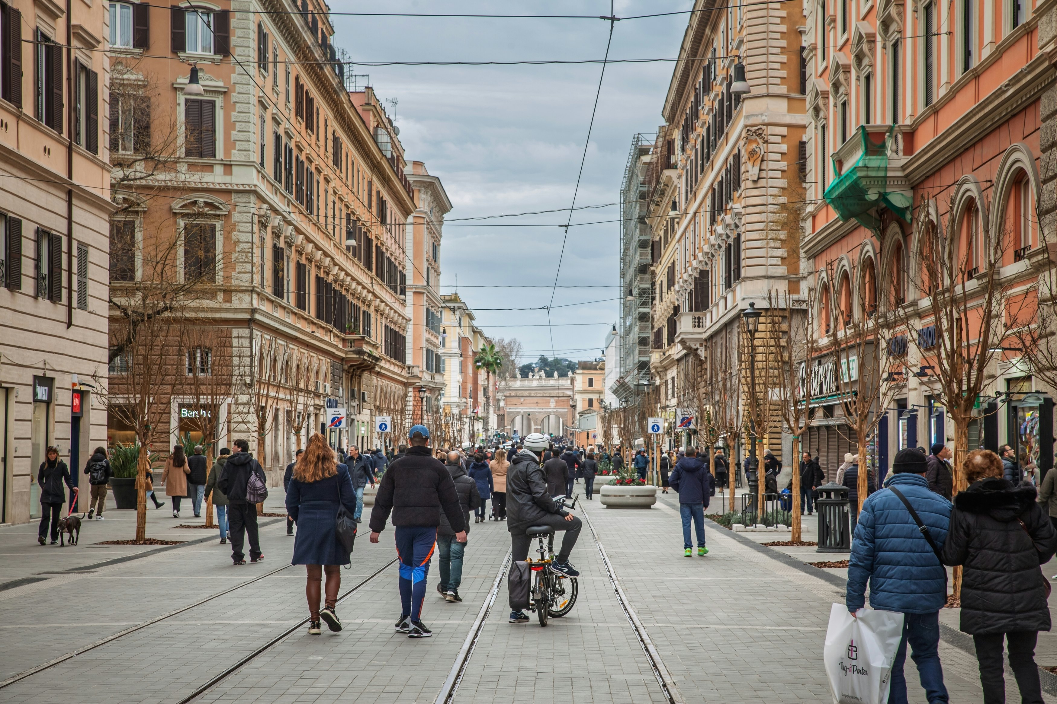 People walking on a busy street with cyclists and tram lines. Historic facades and shops frame pedestrians.