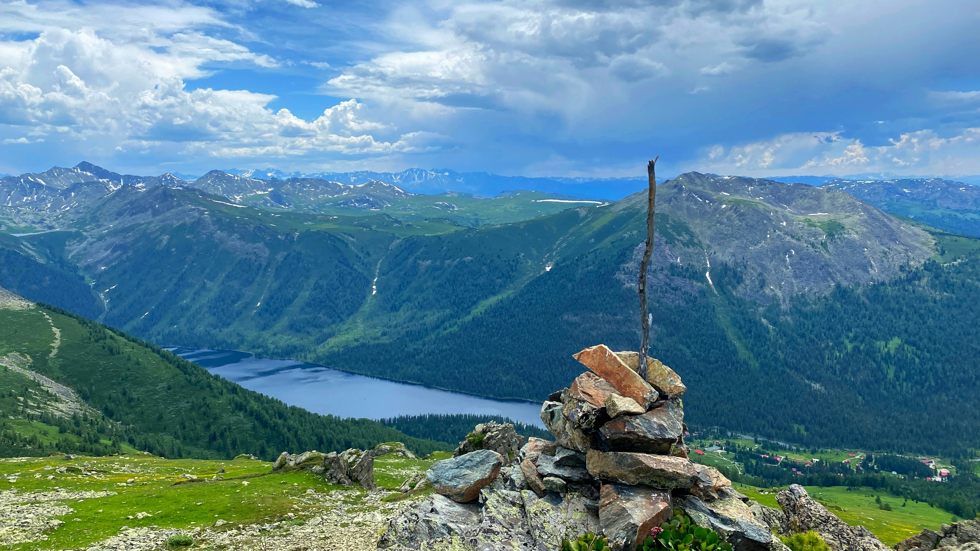 A cairn with a stick at the top stands at an viewpoint overlooking an alpine lake and mountains in the distance.