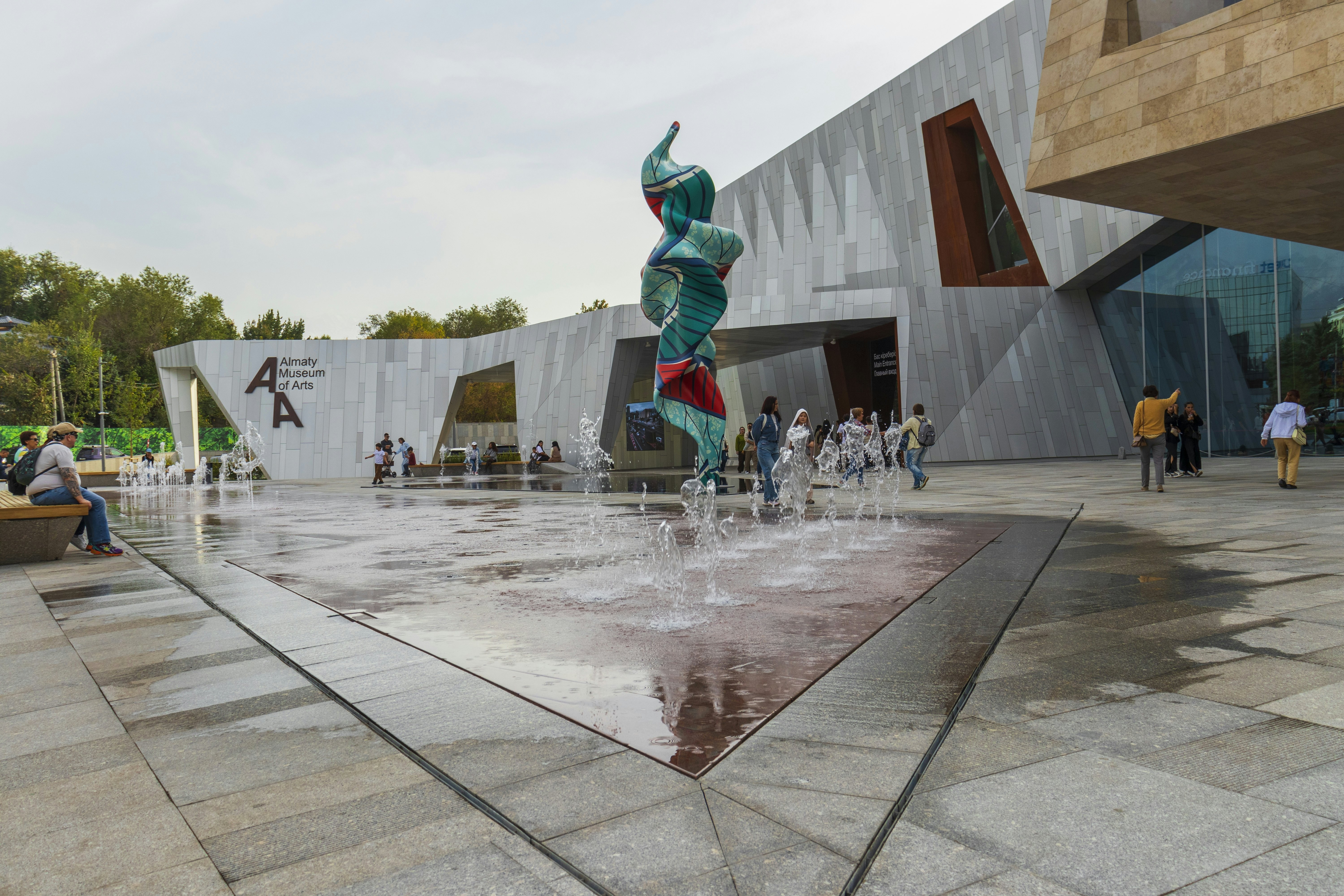 A plaza with a water feature with a sculpture at is center is in front of a museum with contemporary architecture.