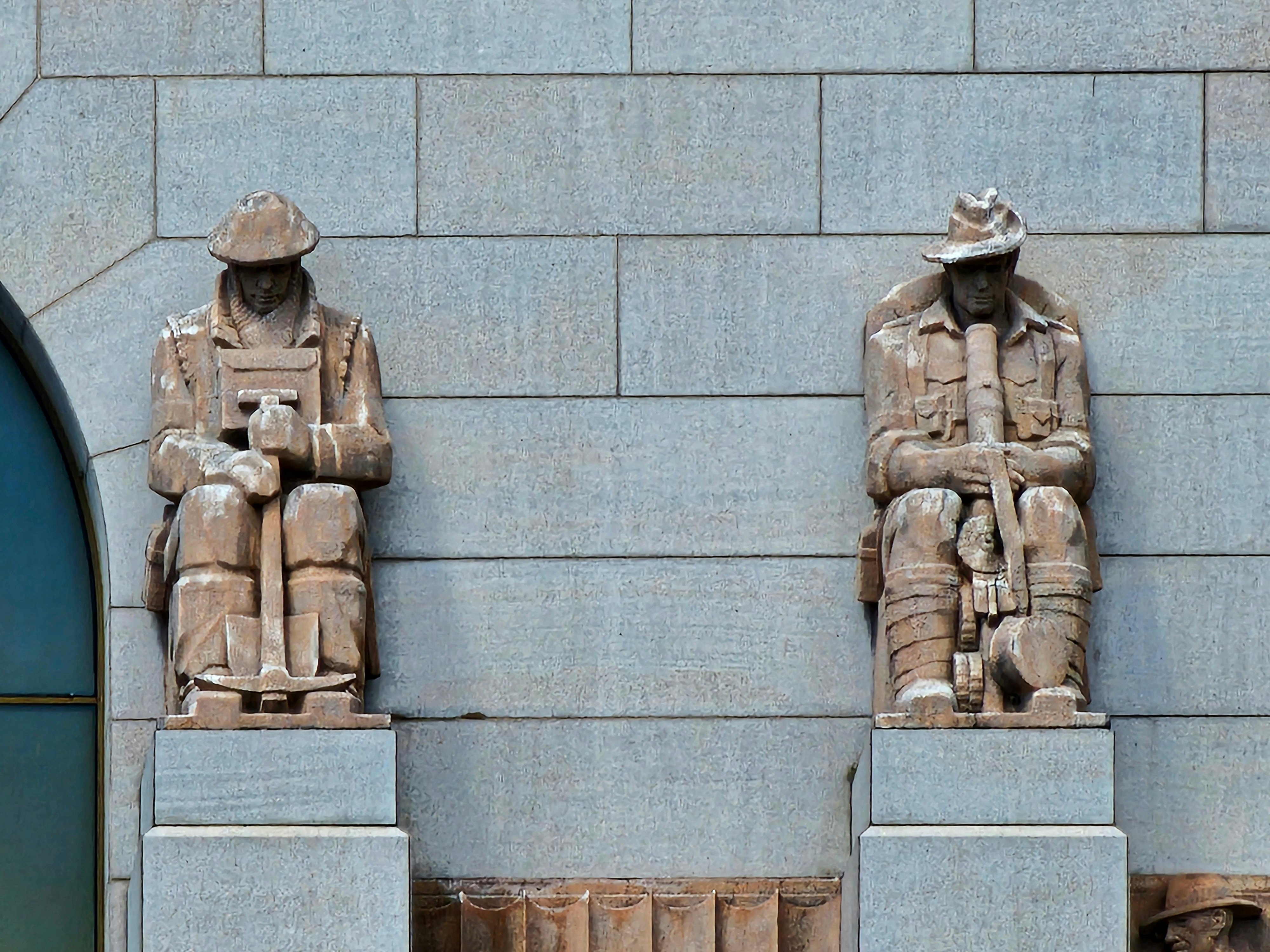Two sculptures of soldiers stand on the exterior of a war memorial in a city.