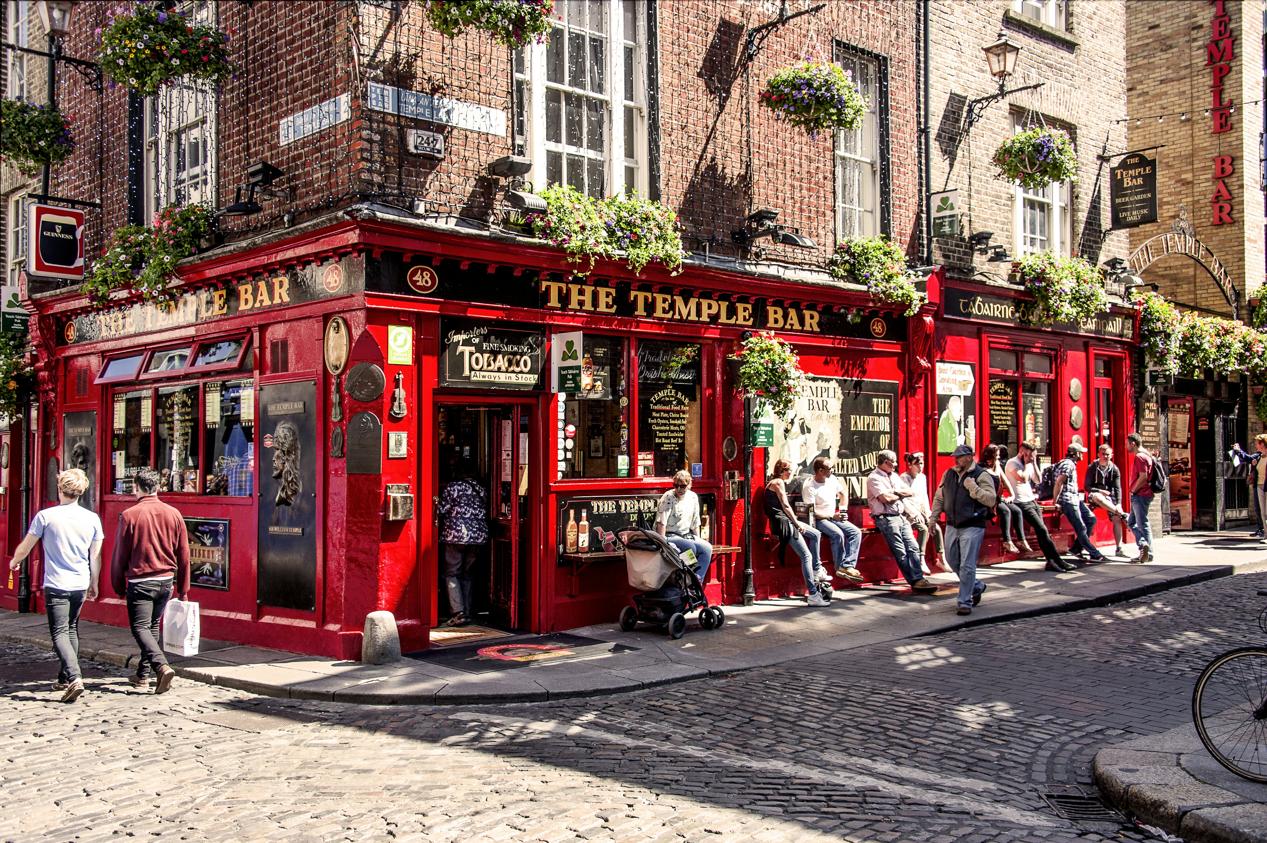 People walking along a cobblestone streeet and sitting outside a corner bar with a bright red facade with hand lettering above the doorway reading "The Temple Bar" on a sunny day.