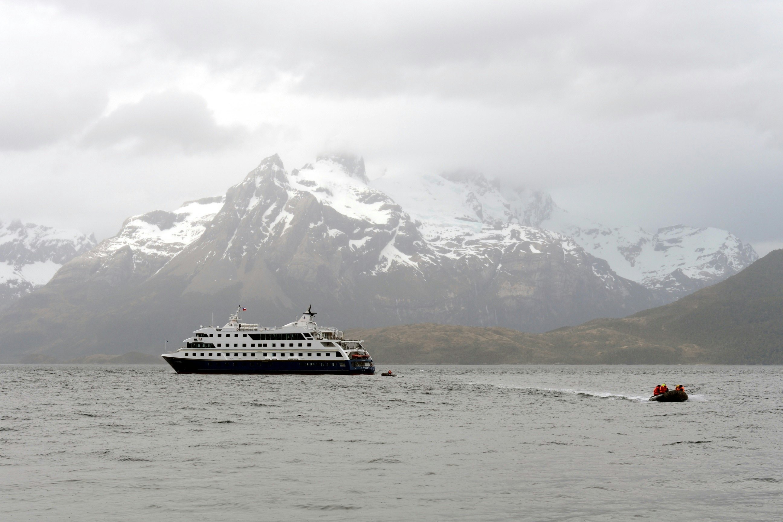 A tender near a larger boat in a waterway surrounded by misty snow-capped peaks.