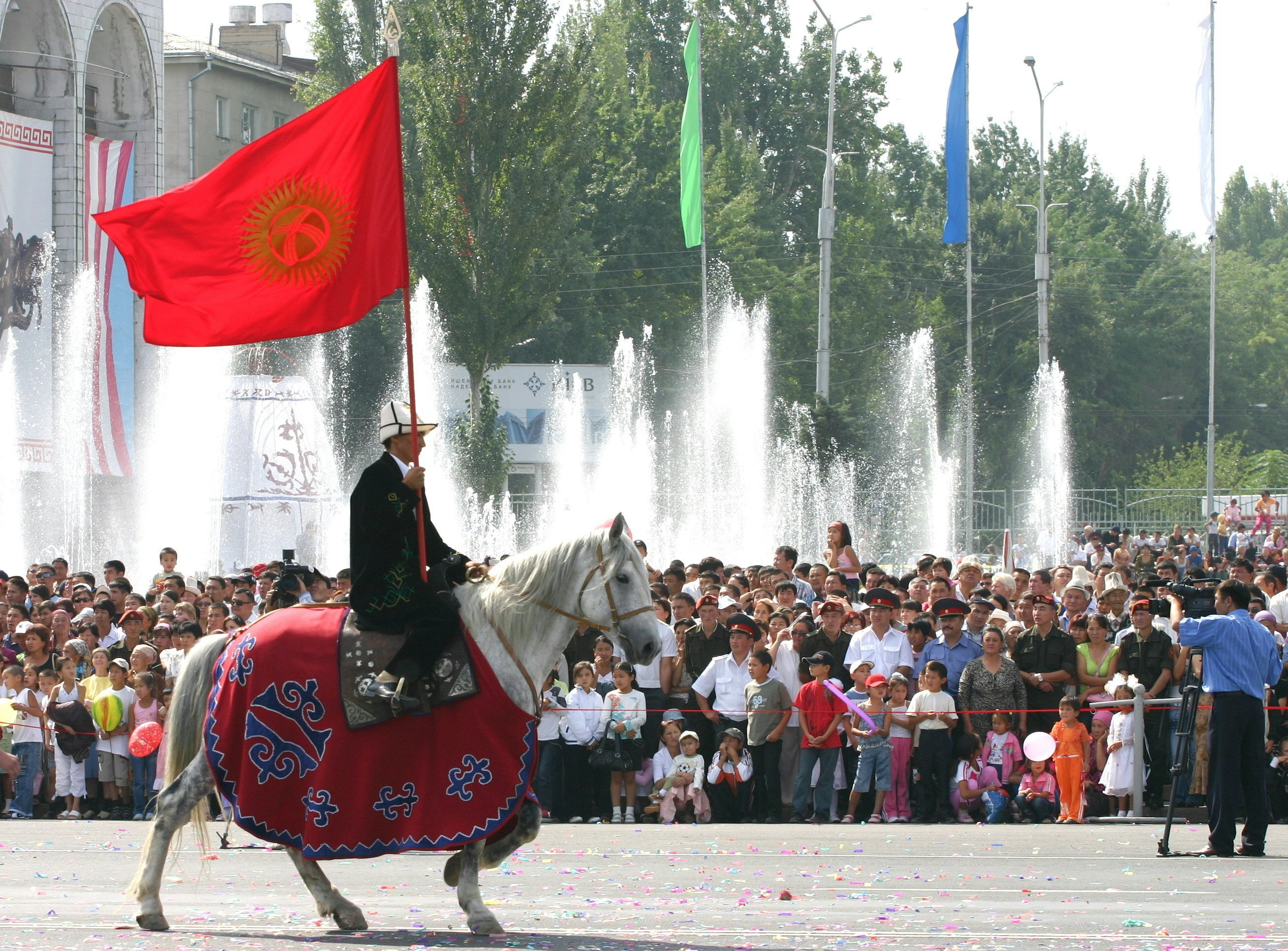 A man carrying the large red flag of Kyrgyzstan rides horseback in a city square for Independence Day celebrations as a crowd looks on.