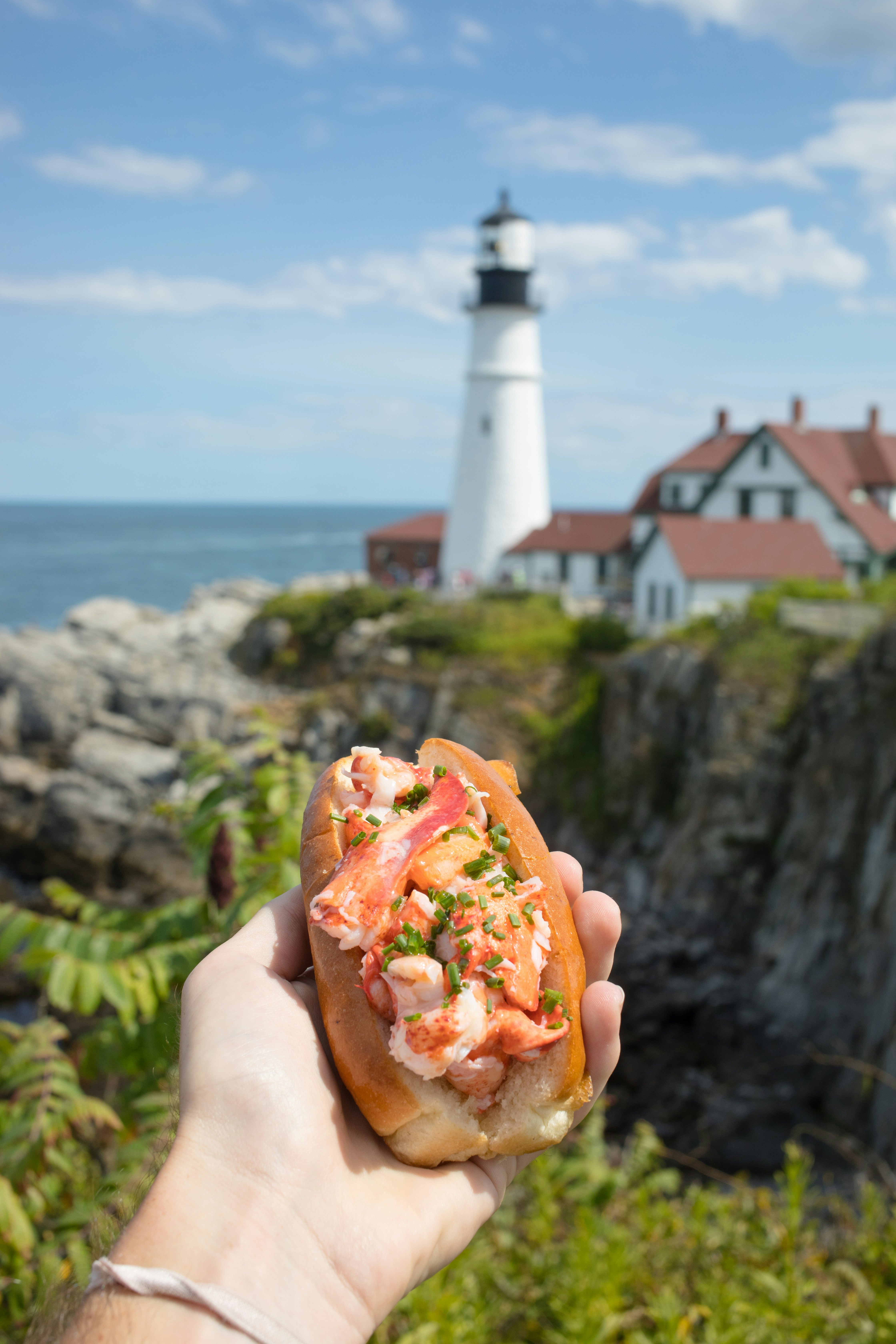 A hand holds out a bread roll stuffed with lobster in a coastal setting near a lighthouse.
