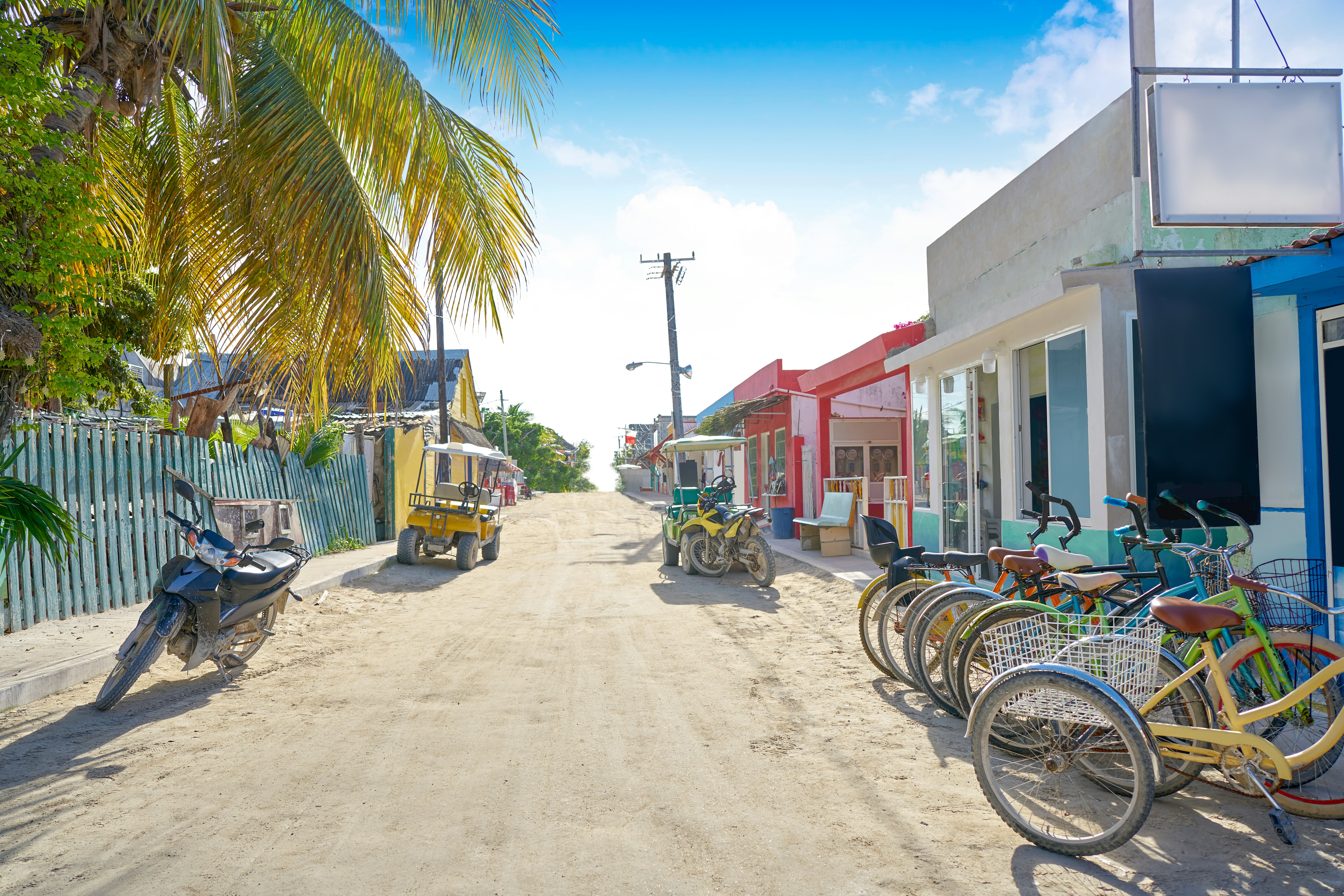 A sandy street in a tropical village is lined with parked bicycles and electric buggies.