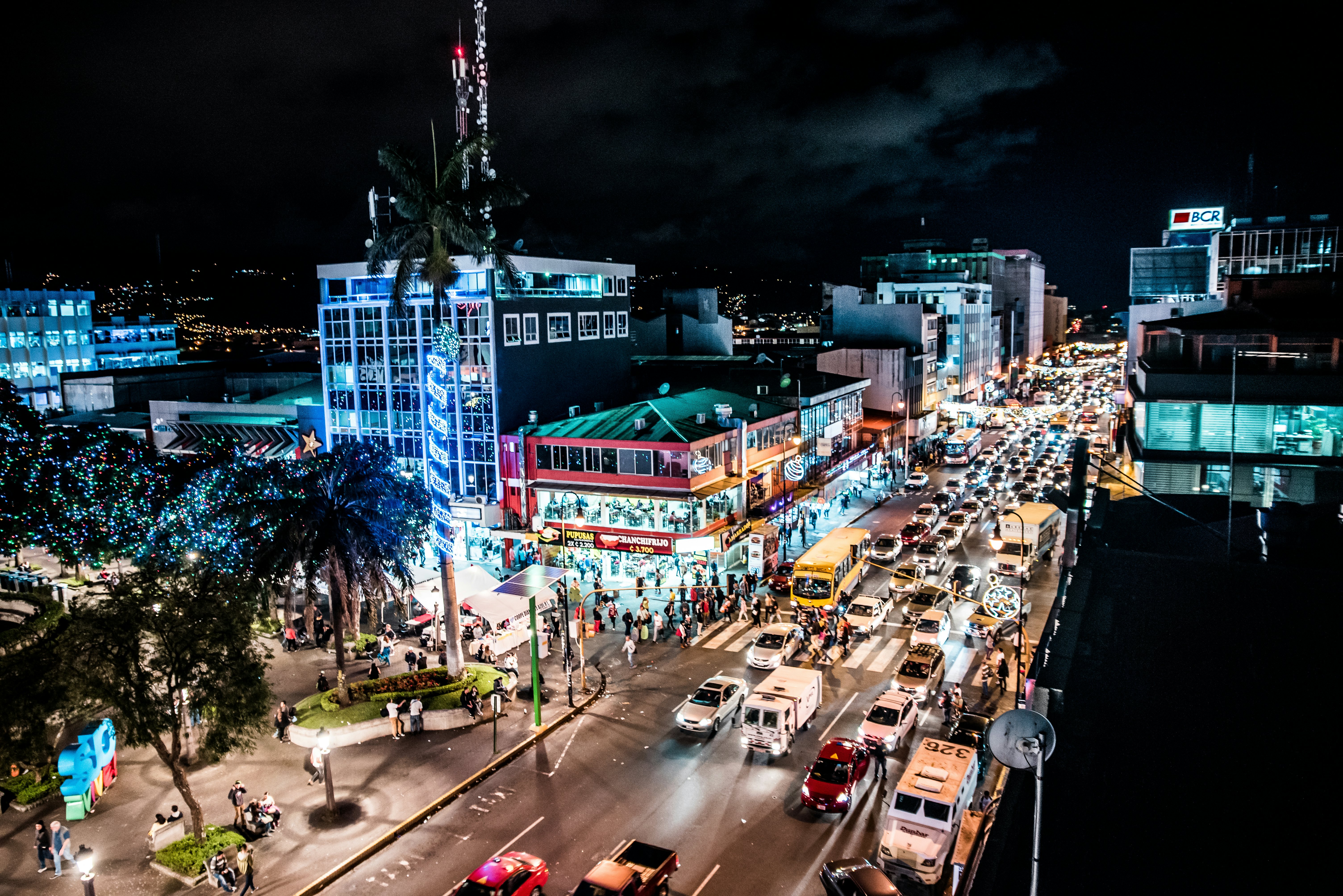 An overview of traffic on a busy street in a city at night. Illuminated buildings line the street and people walk on the sidewalks.