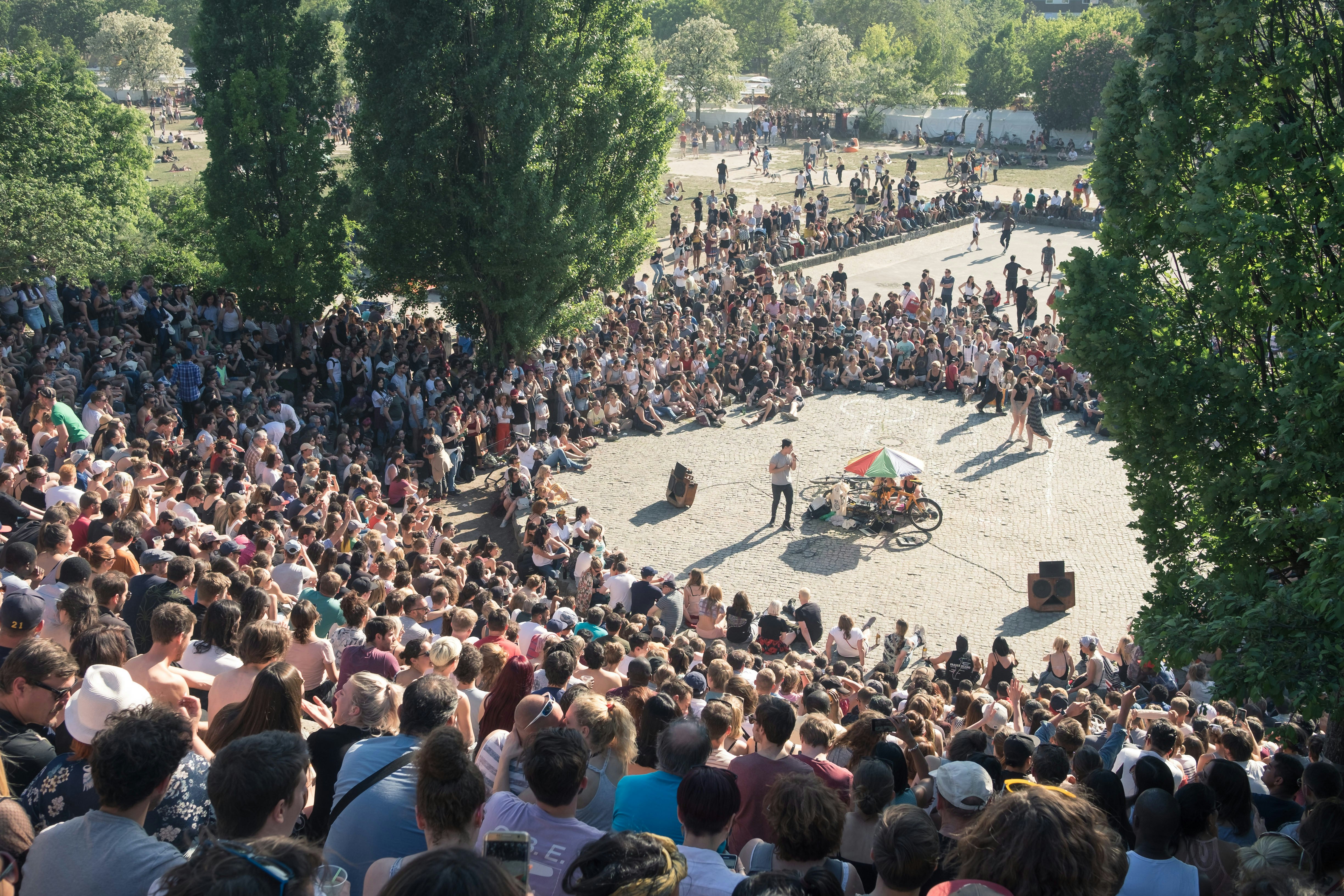 Thousands of people sit in an amphitheater in a city park watching a performer.