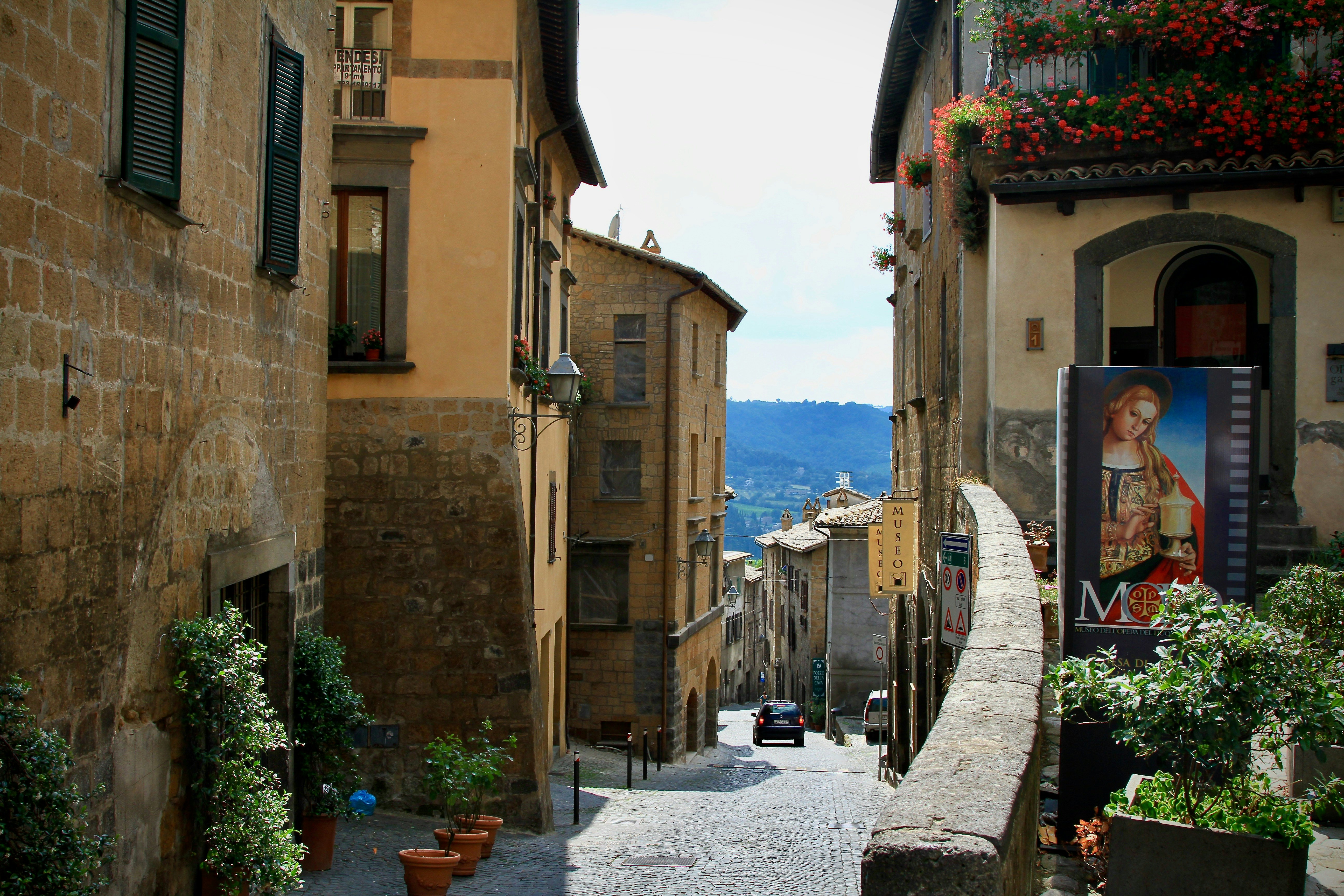 A car moving away from the camera down a narrow cobblestone street lined with earth-toned buildings that have flowers and plants around their doors and windows