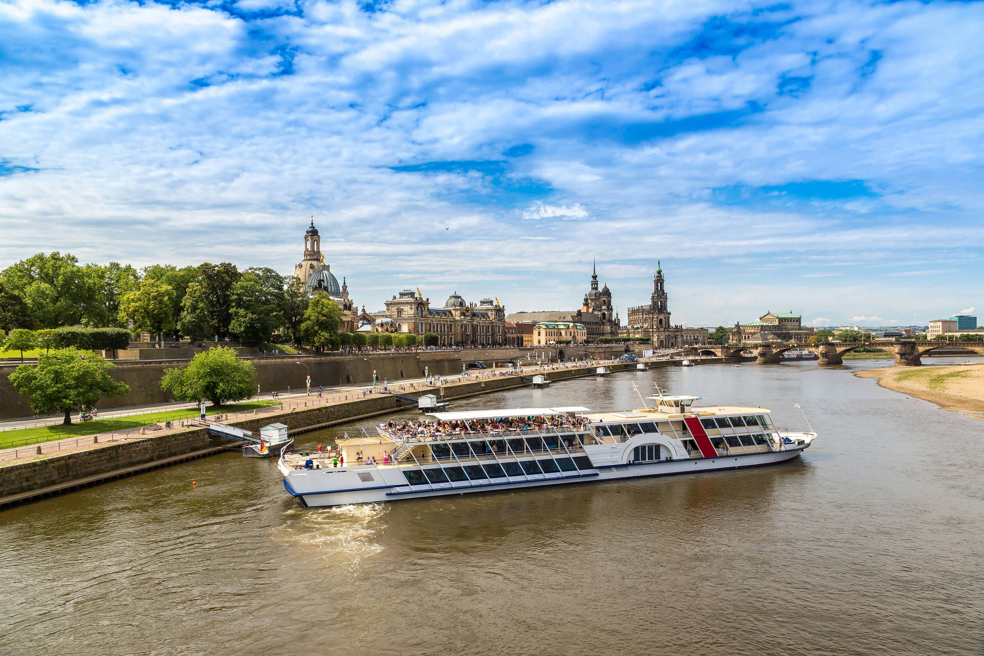 A large passenger ferry in a somewhat narrow river with a city skyline of old, stone buildings in the distance on a sunny day.
