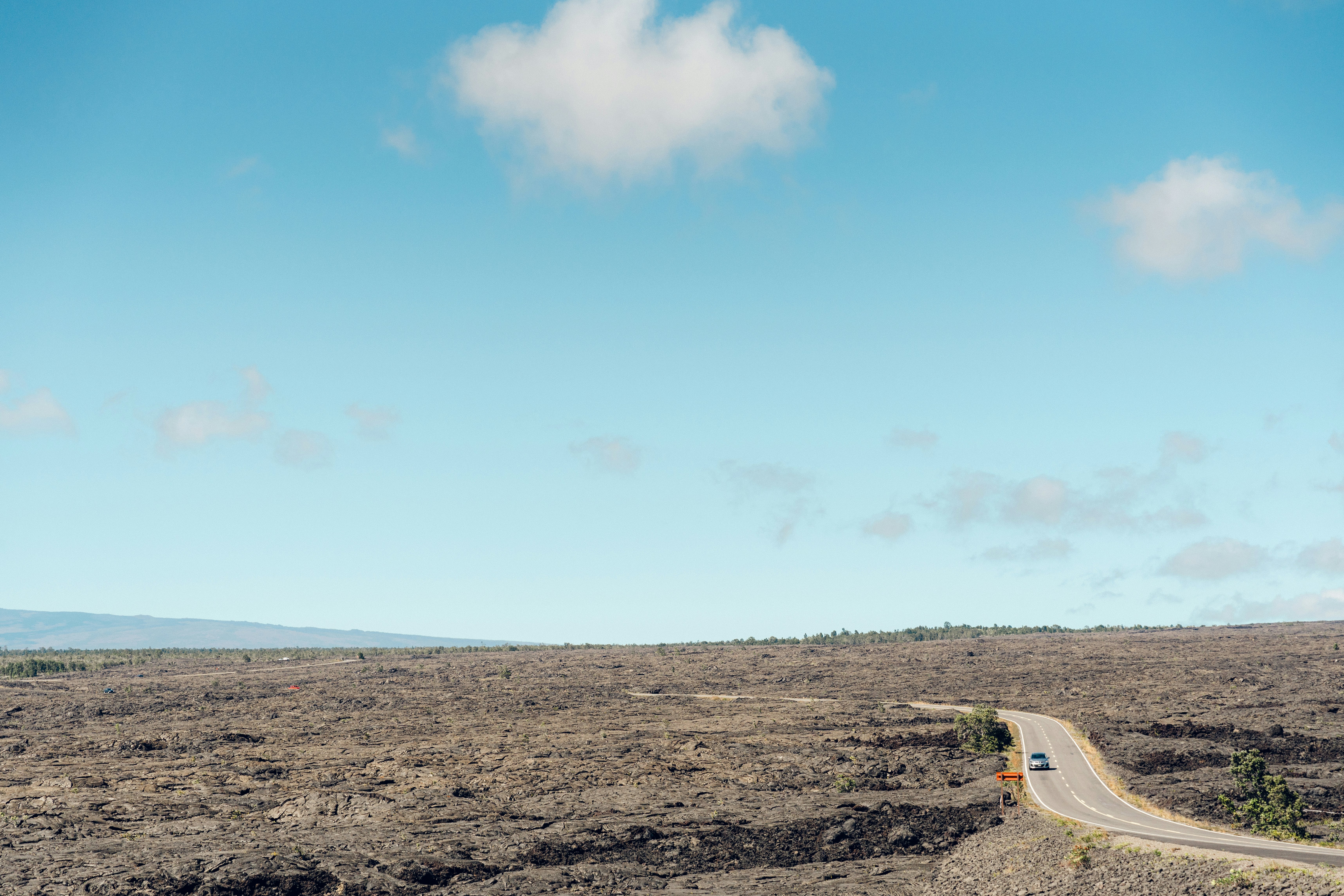 A car travels on a two-lane road through a barren volcanic landscape on a sunny day.