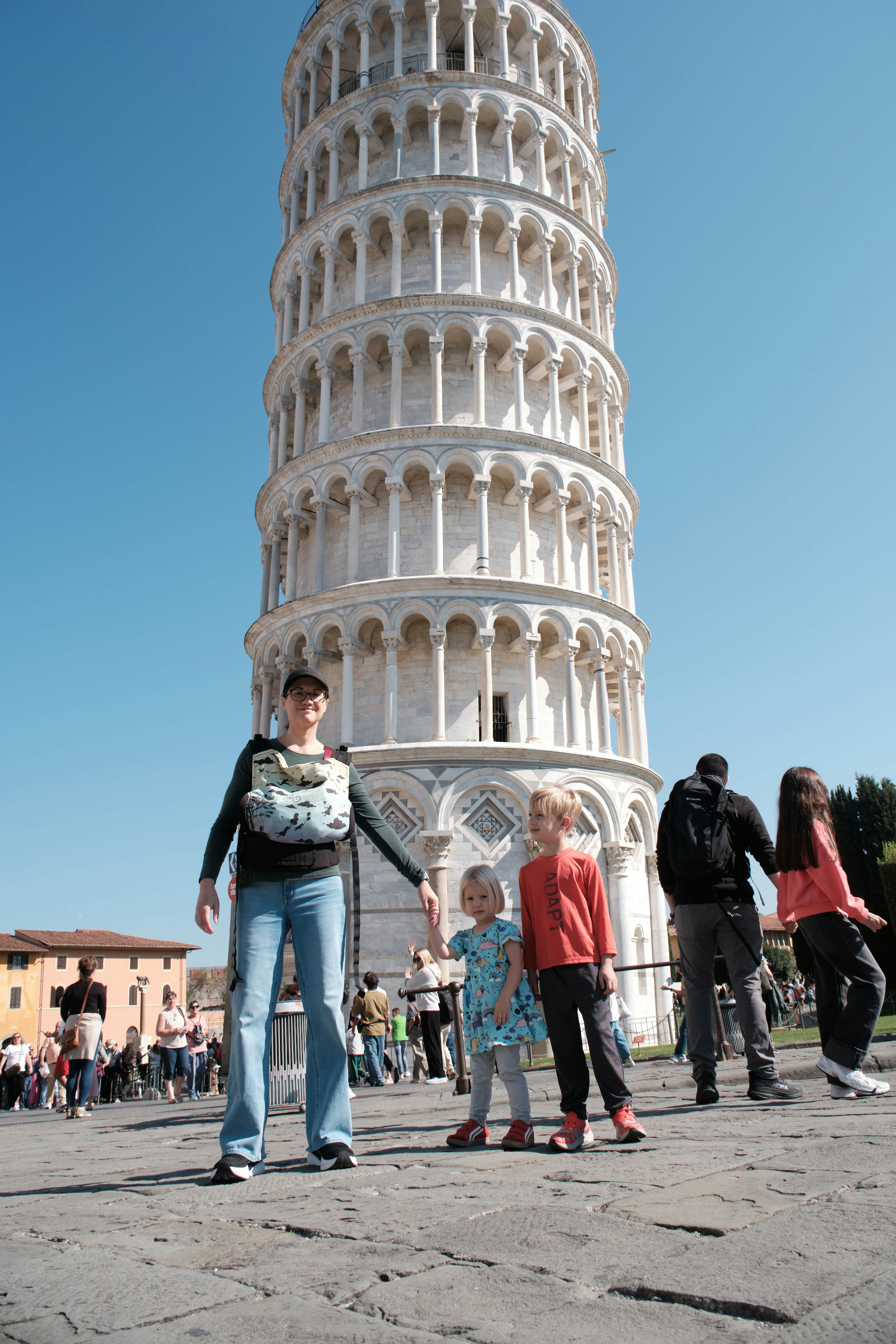 A woman and two small children stand in front of an ornate round tower that stands at an angle.