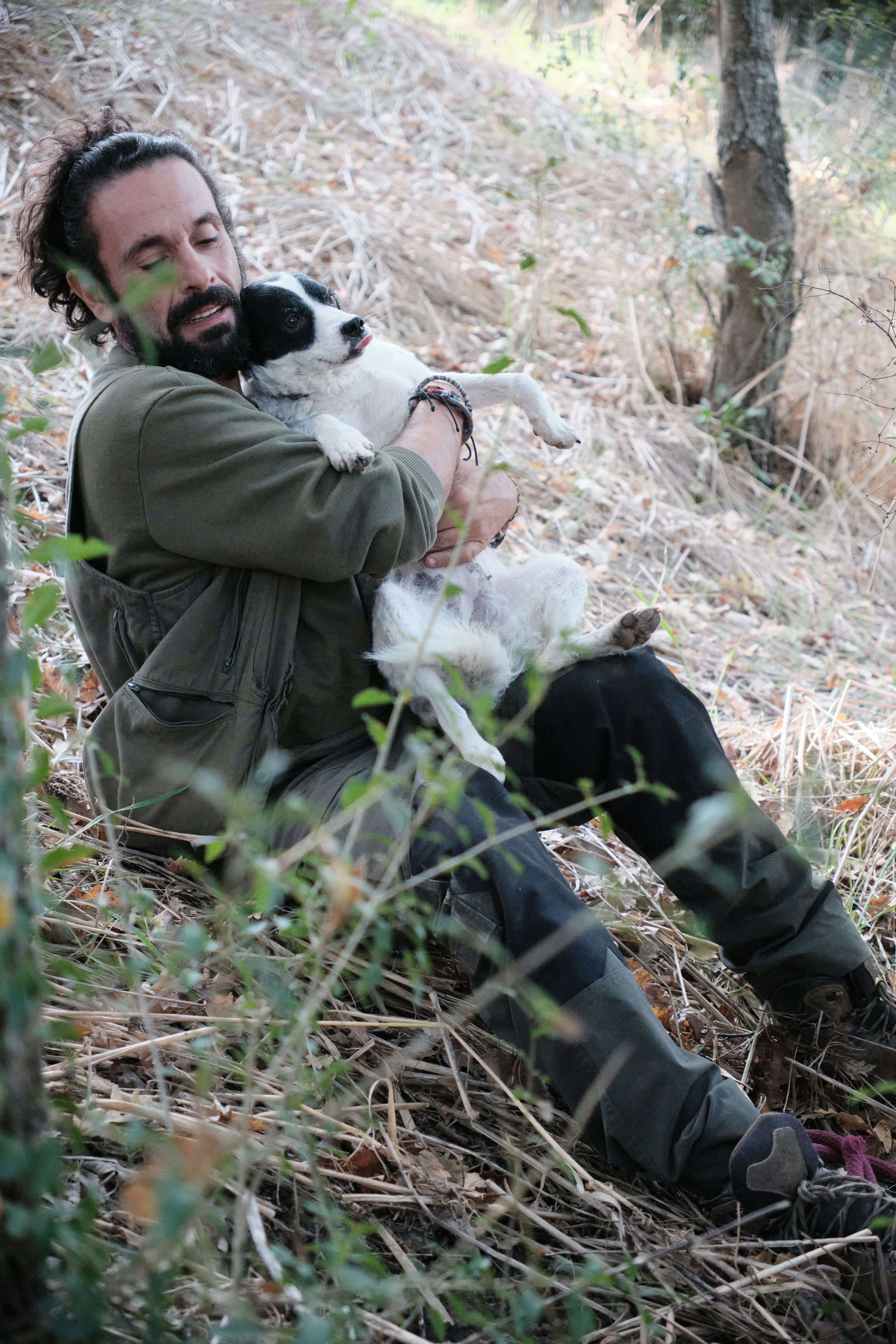 A man sits on the grass in woodland cuddling a wriggling black and white dog.