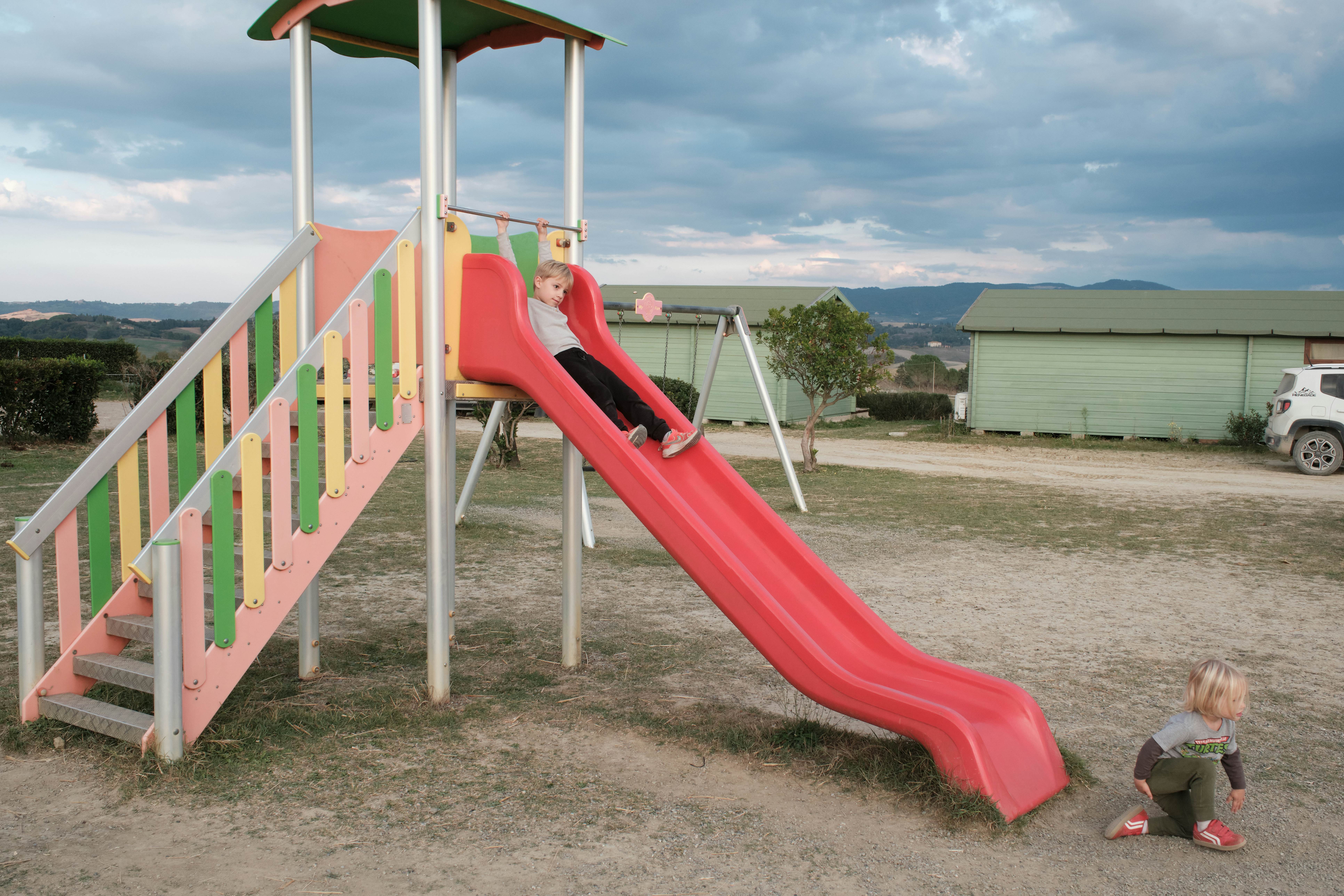 A play area with a slide. One child hangs on the top as another clamber up from the ground below.