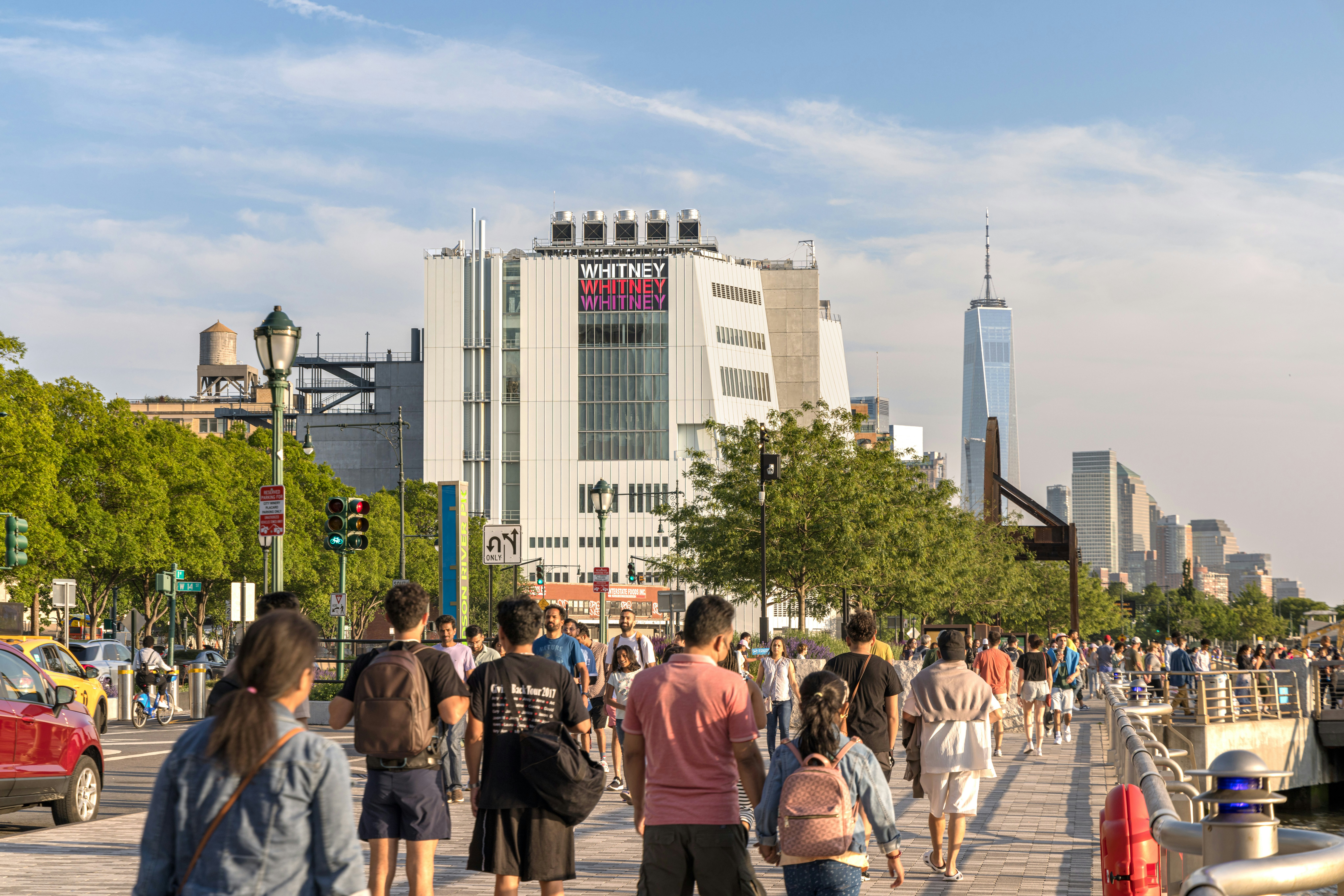 People walk in a riverfront park in a big city. A large museum is seen across the road.
