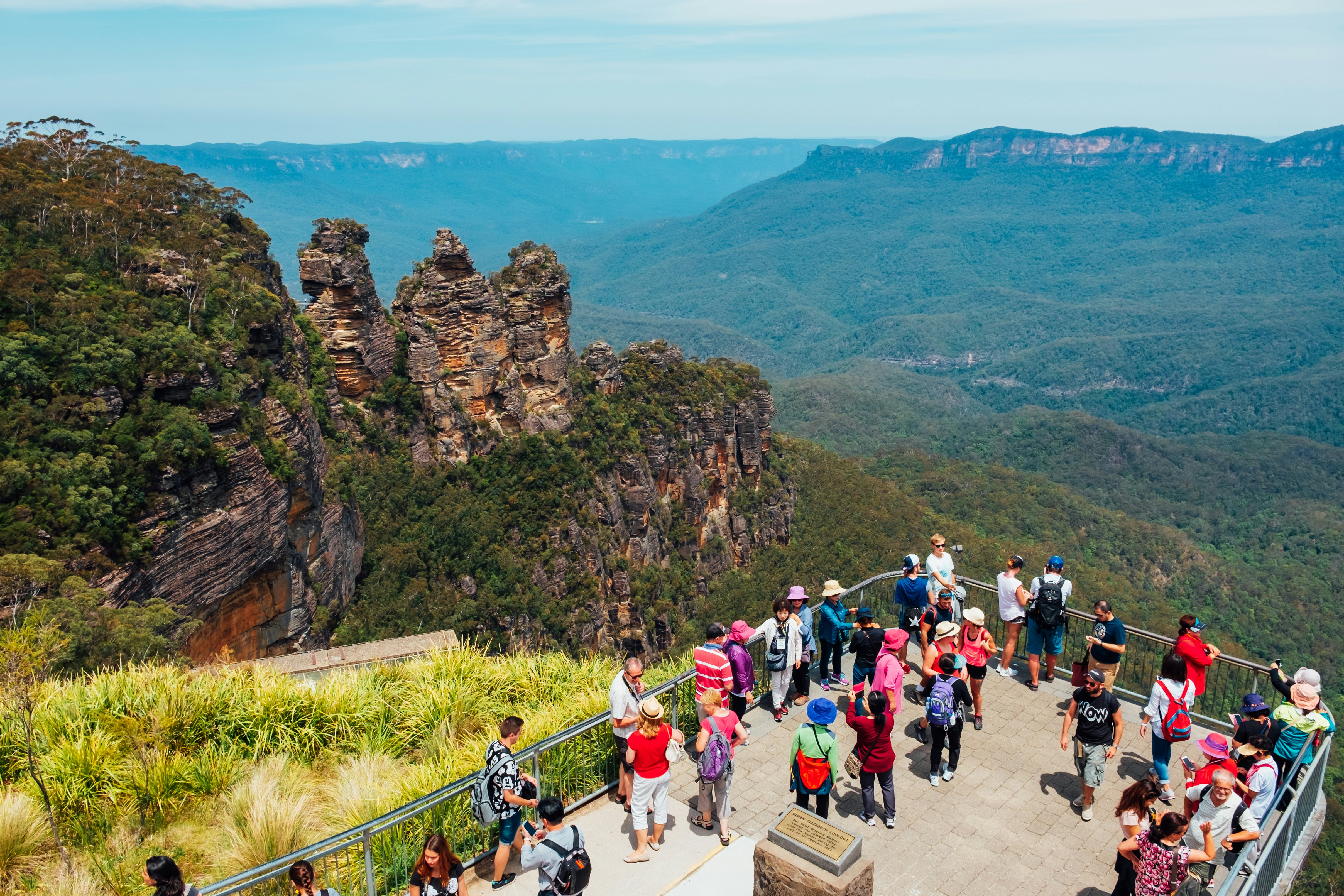 A group of tourists at a lookout point take photos of the scenery. Rolling hills covered in thick forest stretch into the distance. A rock formation of three distinct pillars stands nearby.