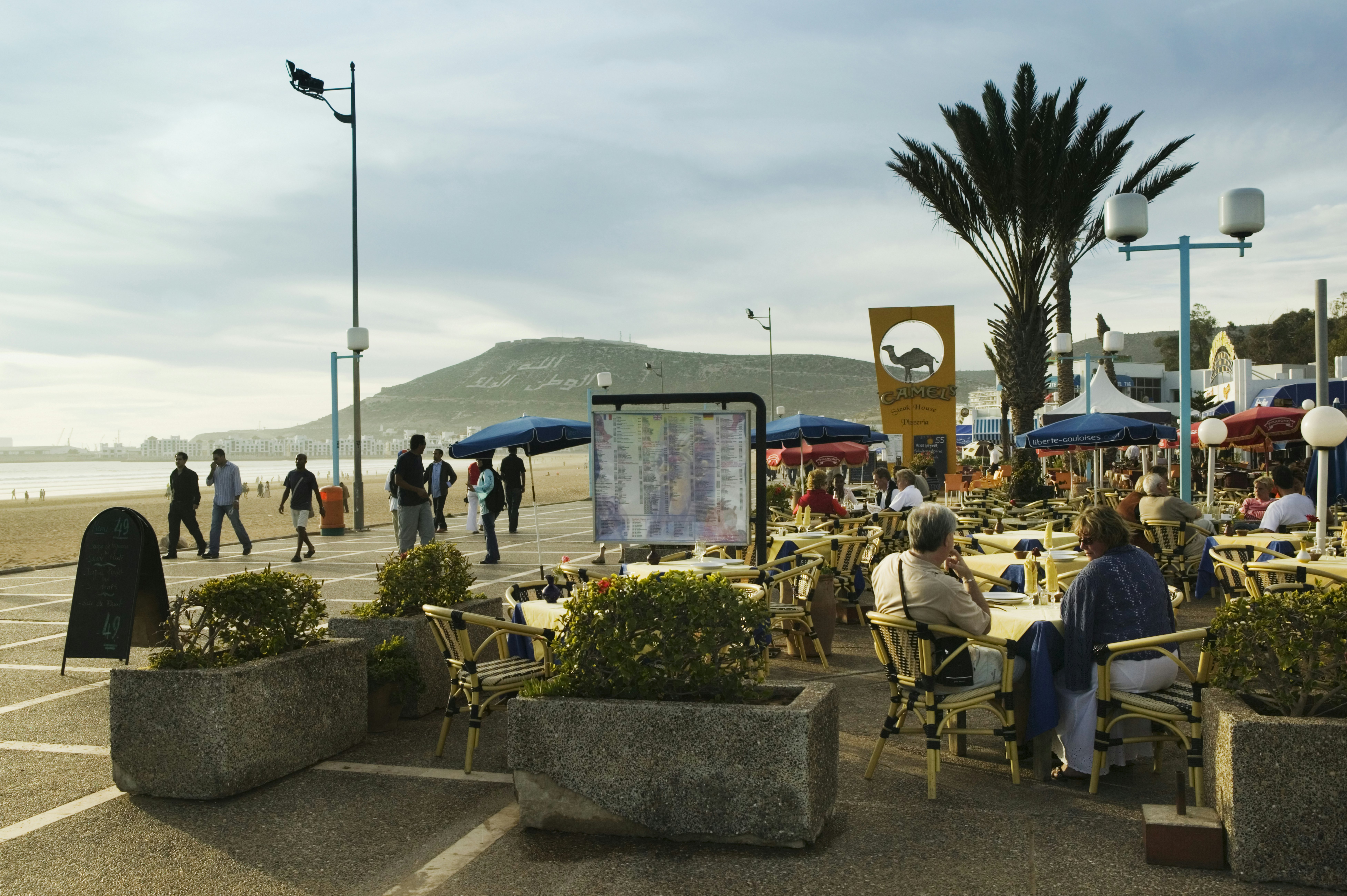 People sit at a cafe and walk along a promenade by a beach in Morocco.