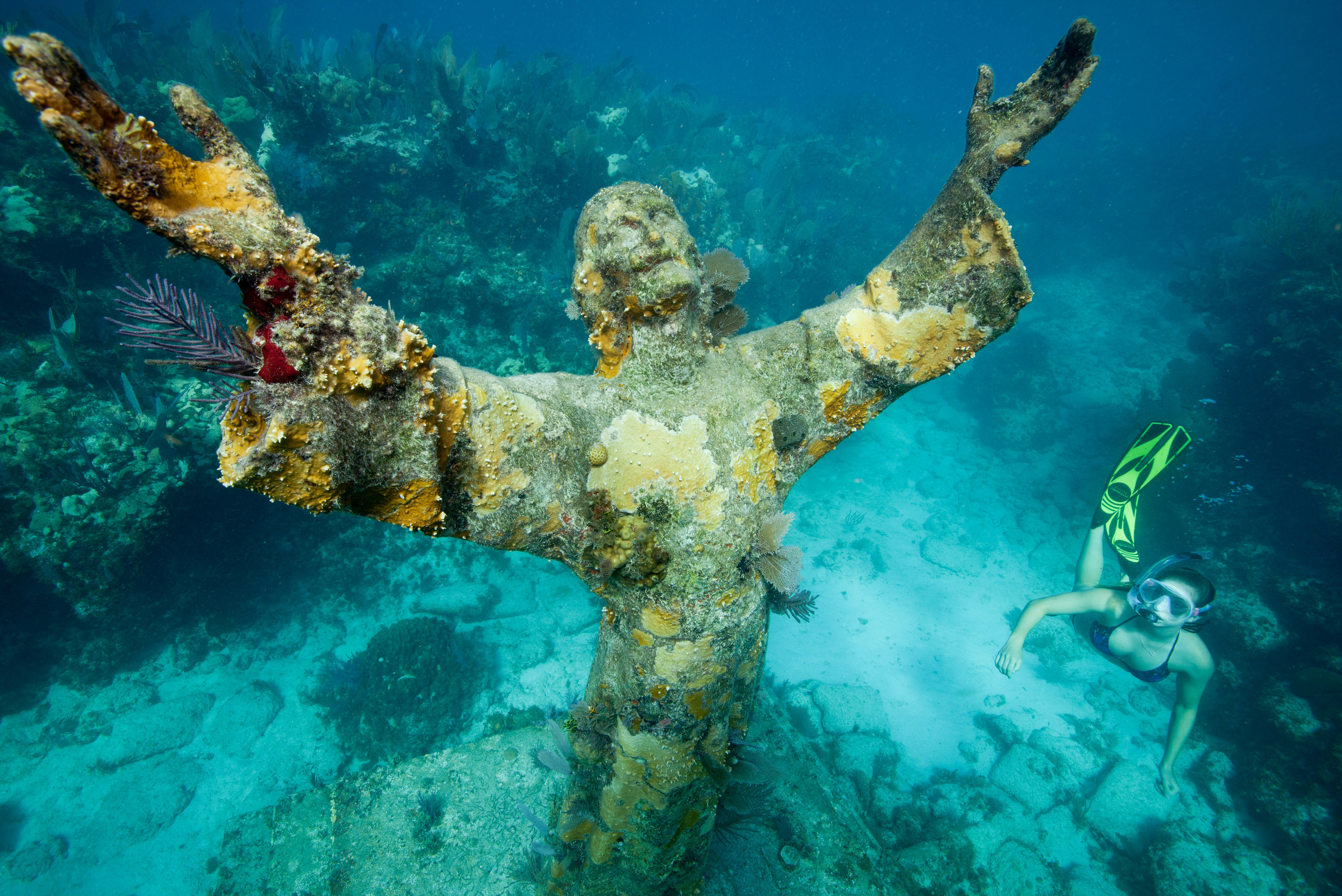 A statue of Jesus Christ sits underwater, encrusted with corals and other marine plants. A snorkeler swims to the right of the statue.