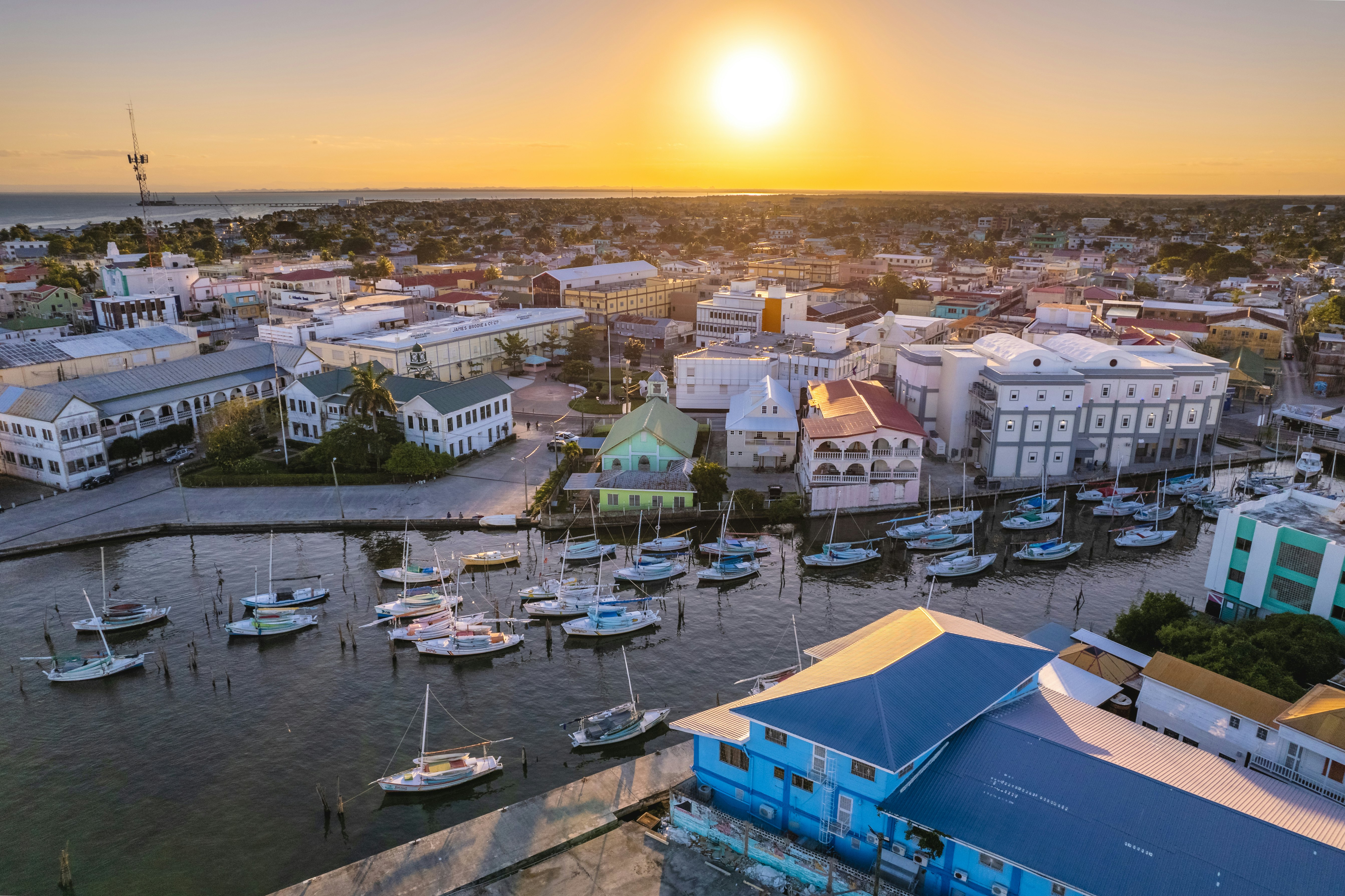 An aerial view of a waterfront in a city at sunset. Buildings with porches line the waterfront, while sailboats are moored in the water.