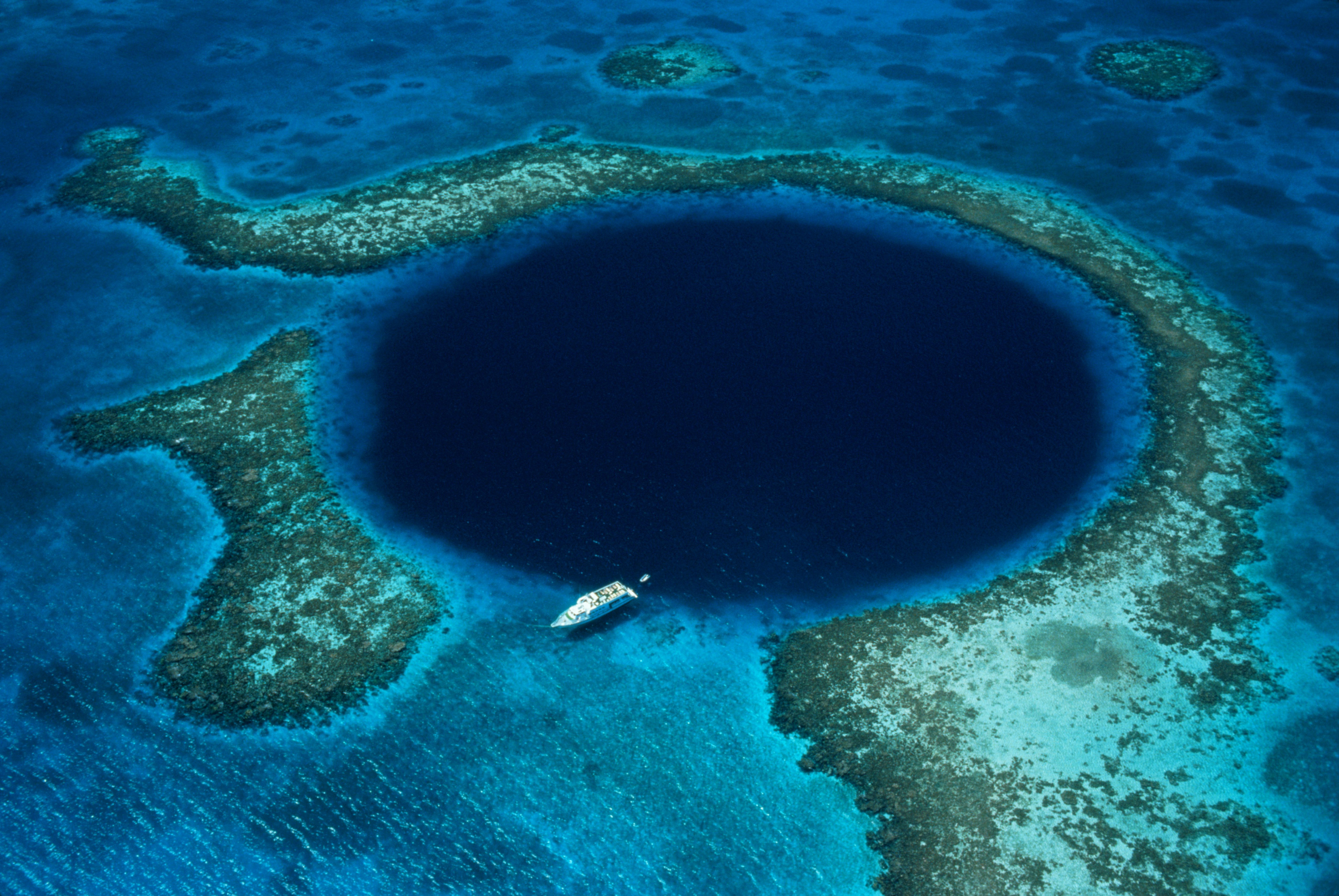 An aerial view of a boat next to the blue waters in a perfectly round sinkhole surrounded by lighter-colored reefs and sand.