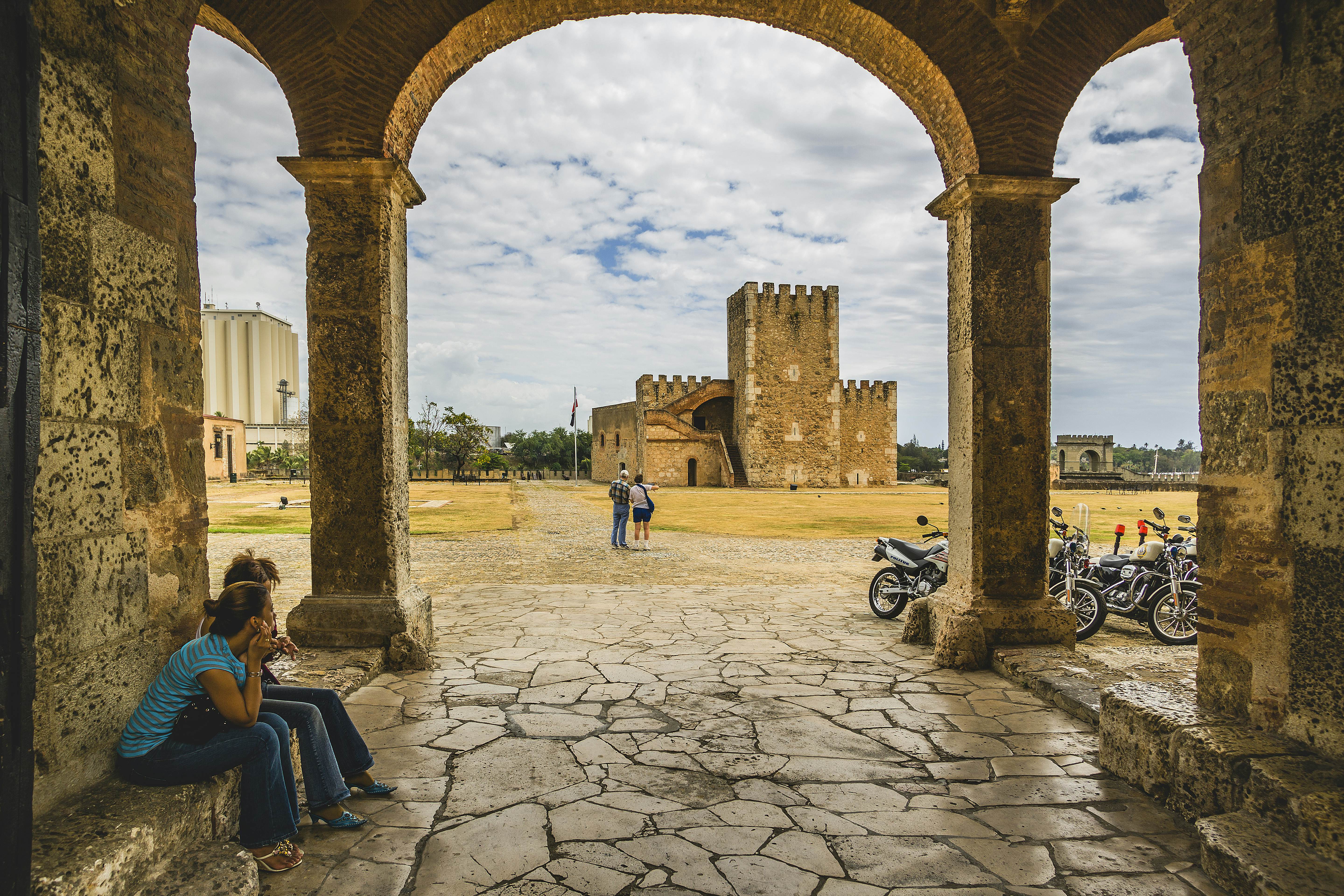A tan stone fort is visible through an archway; two people walk across a long courtyard to reach the fort.