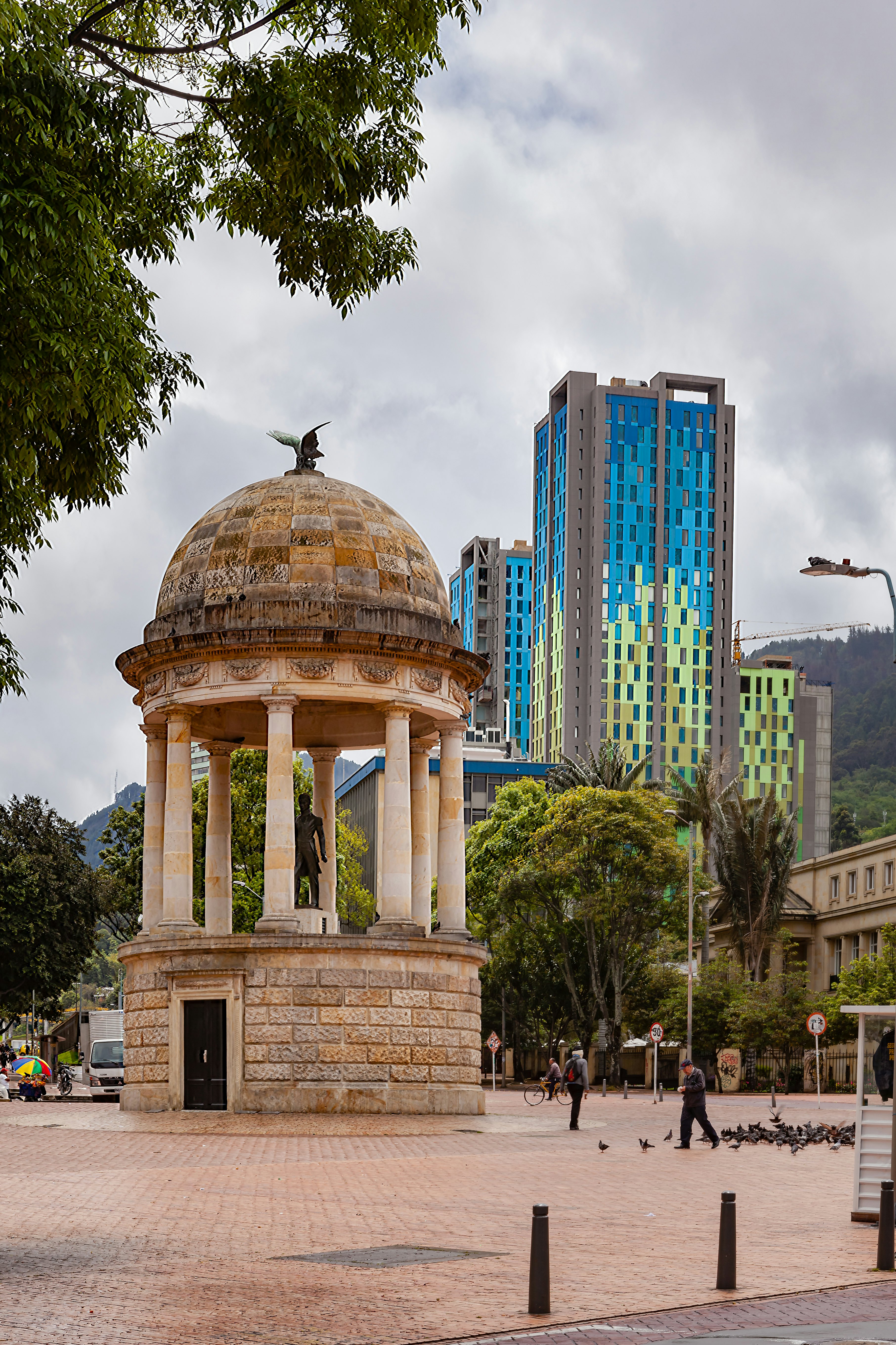 A temple-like monument stands at the center of a city plaza, with a tower seen in the distance.