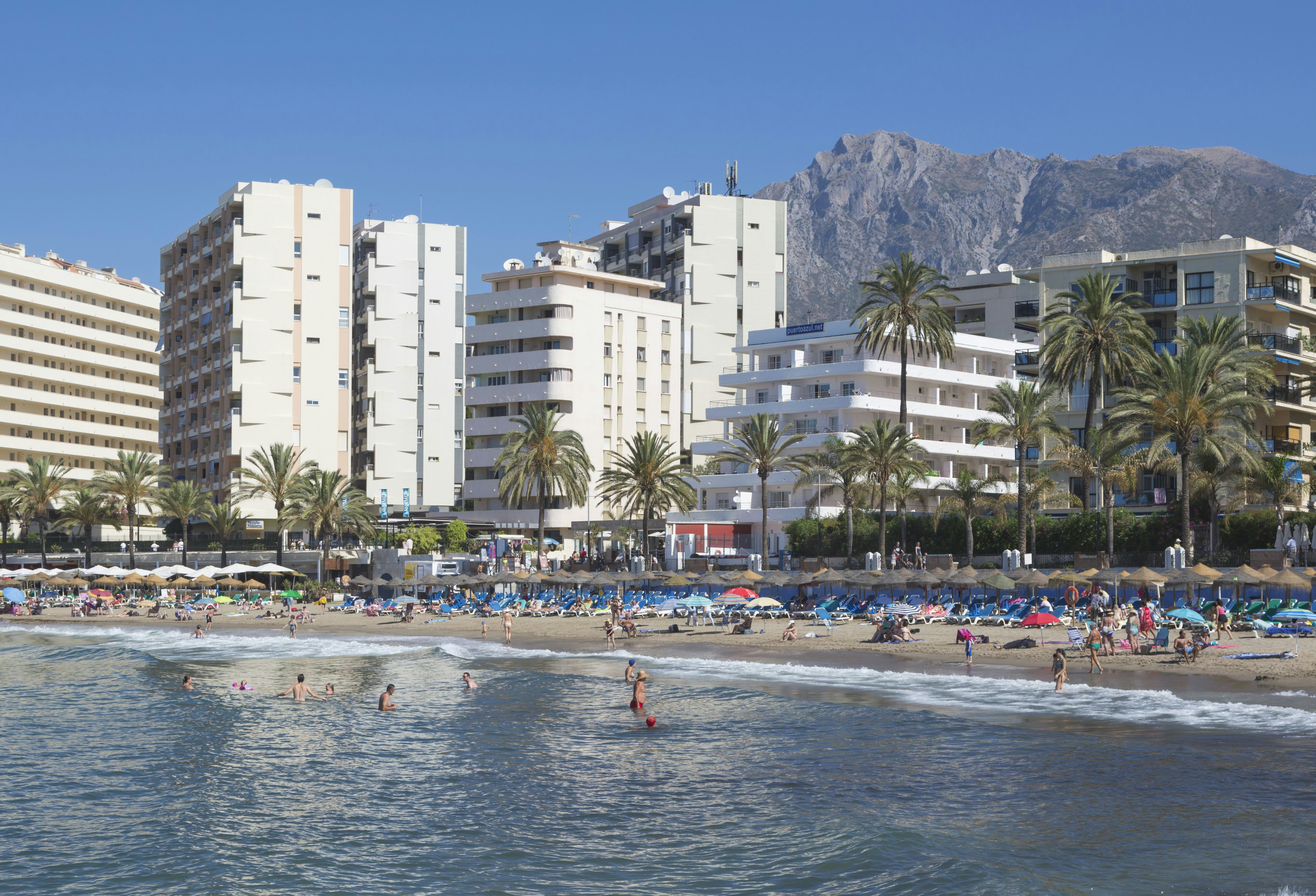People on a beach with blue lounge chairs and in blue water in Spain. Palm trees separate the beach from white midrise buildings behind it; a mountain is in the background.
