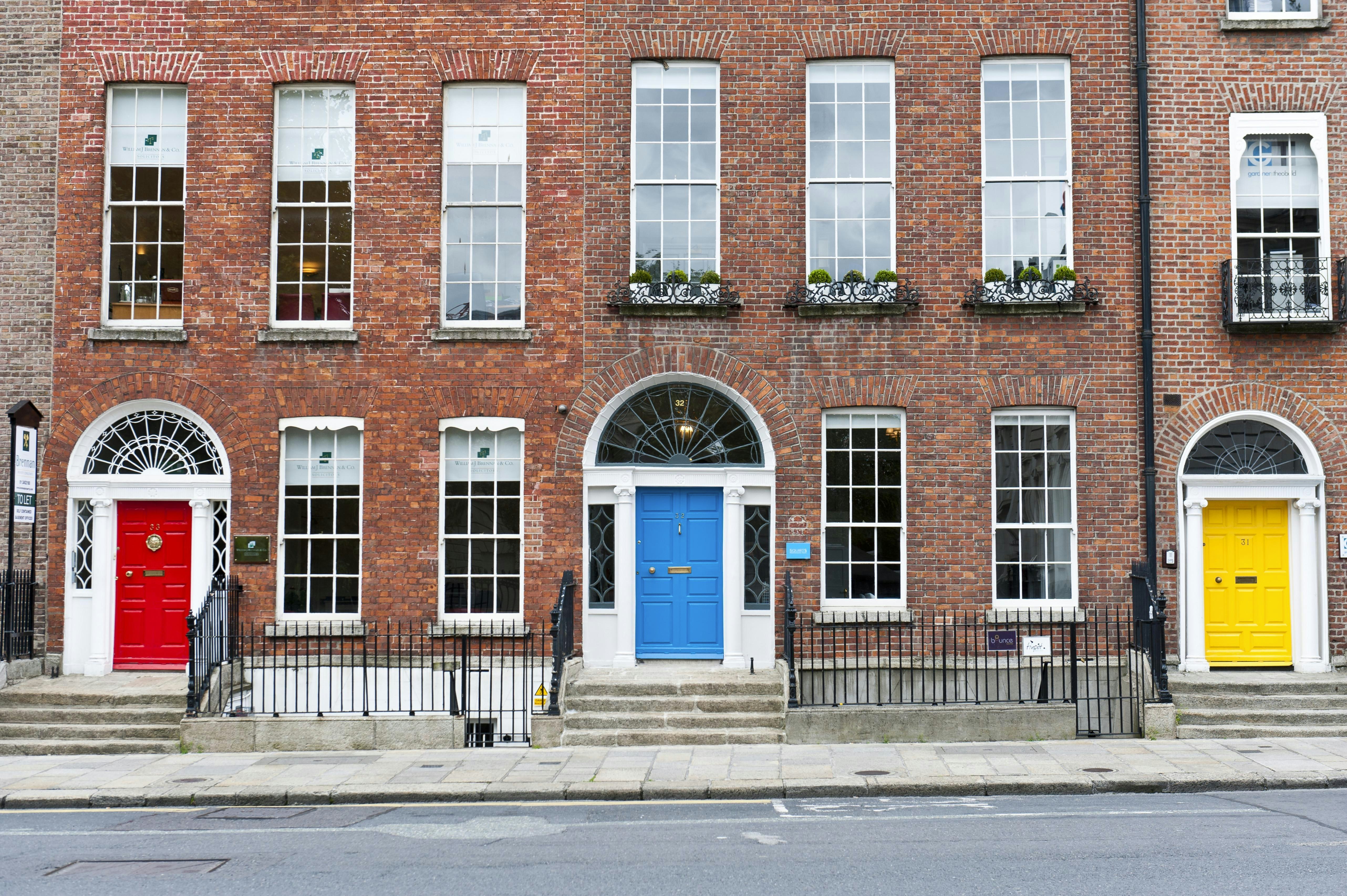 Three brick-fronted buildings are seen on a city street, one with a door painted red, one with a door painted blue and one with a door painted yellow.