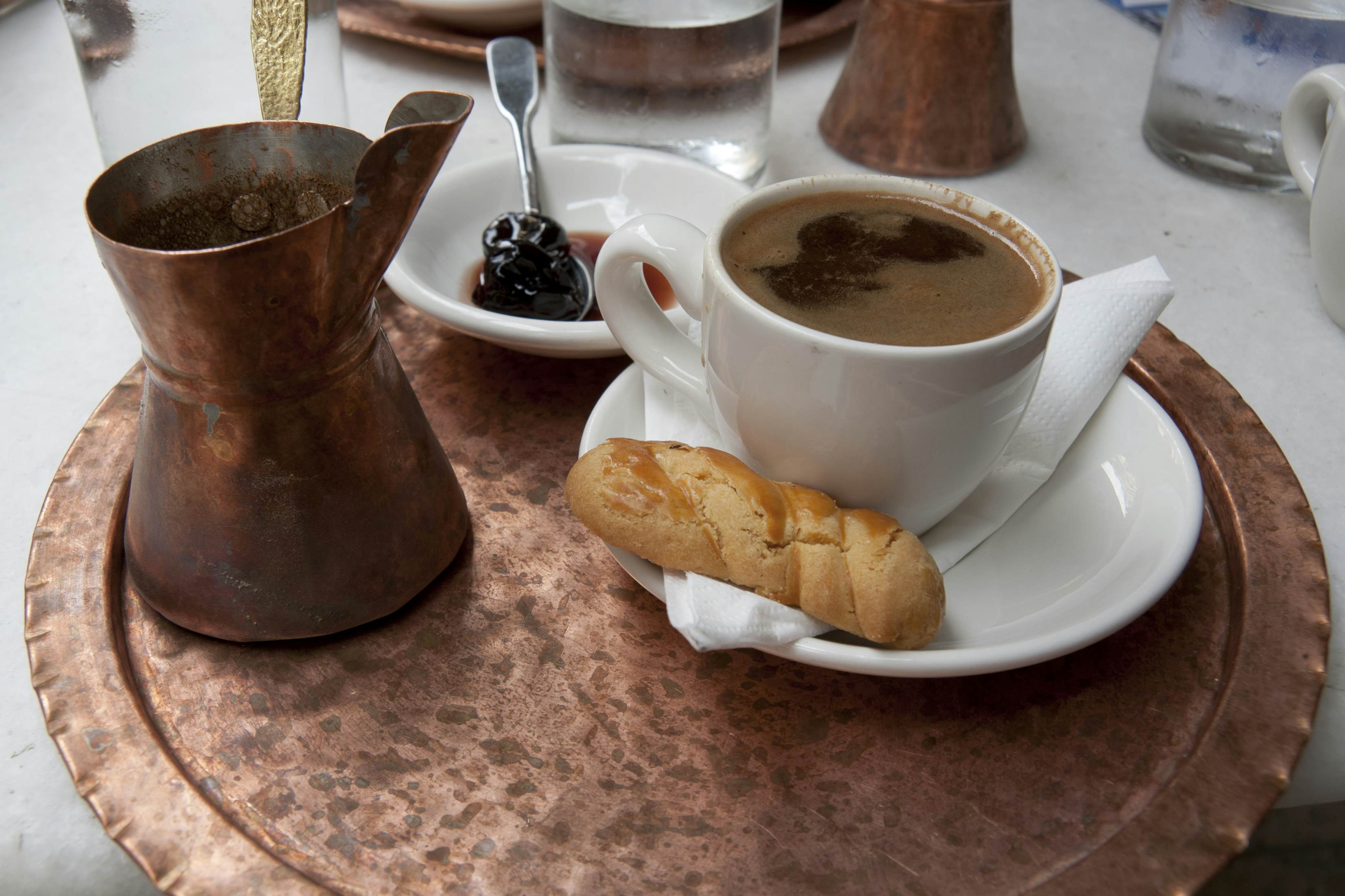 A copper tray with a white cup and saucer with coffee and a cookie, a copper vessel with coffee, and a dish with an item in dark syrup and a spoon.