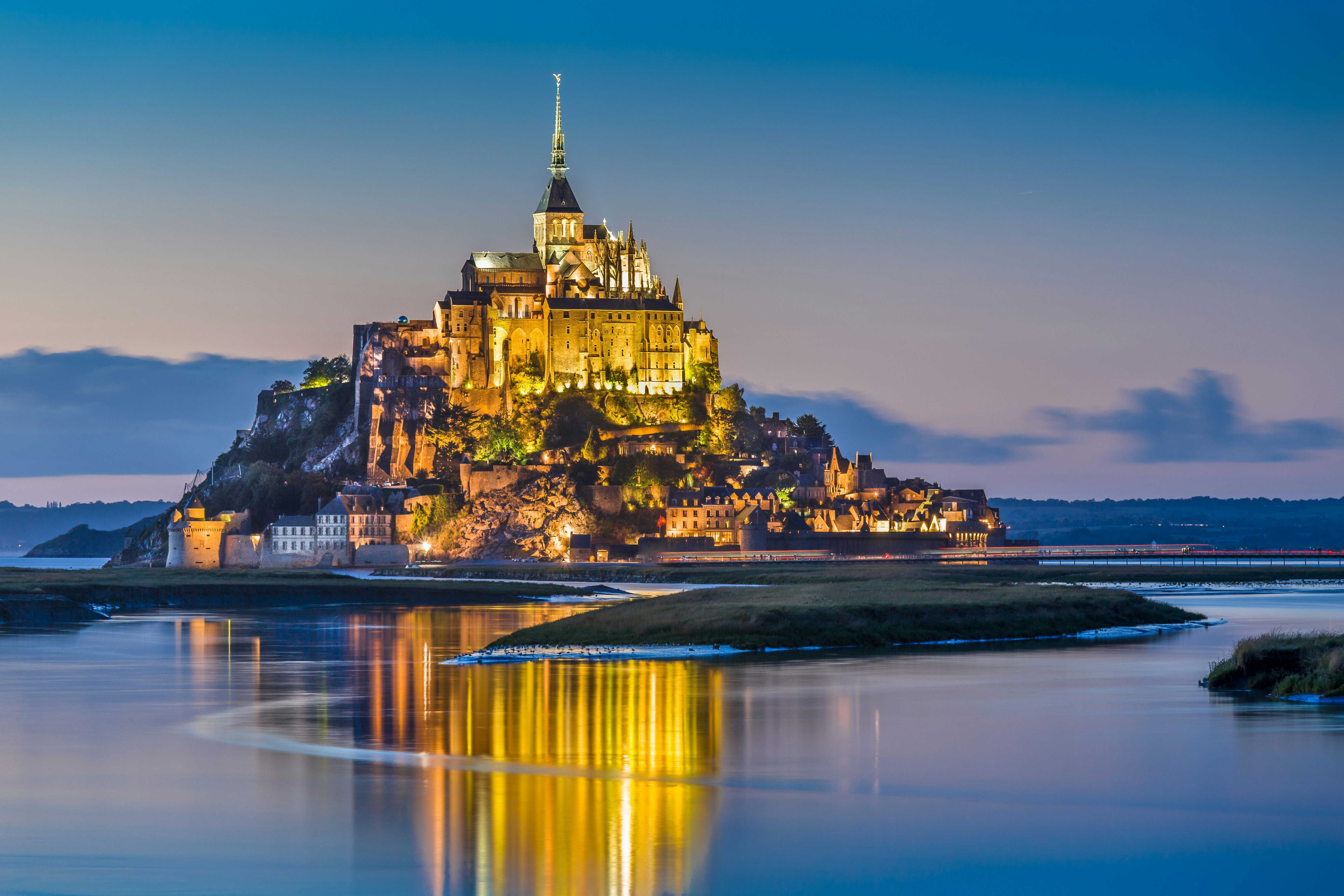 An islet crowned by a monastery stands tall on the sand at low tide in the twilight.