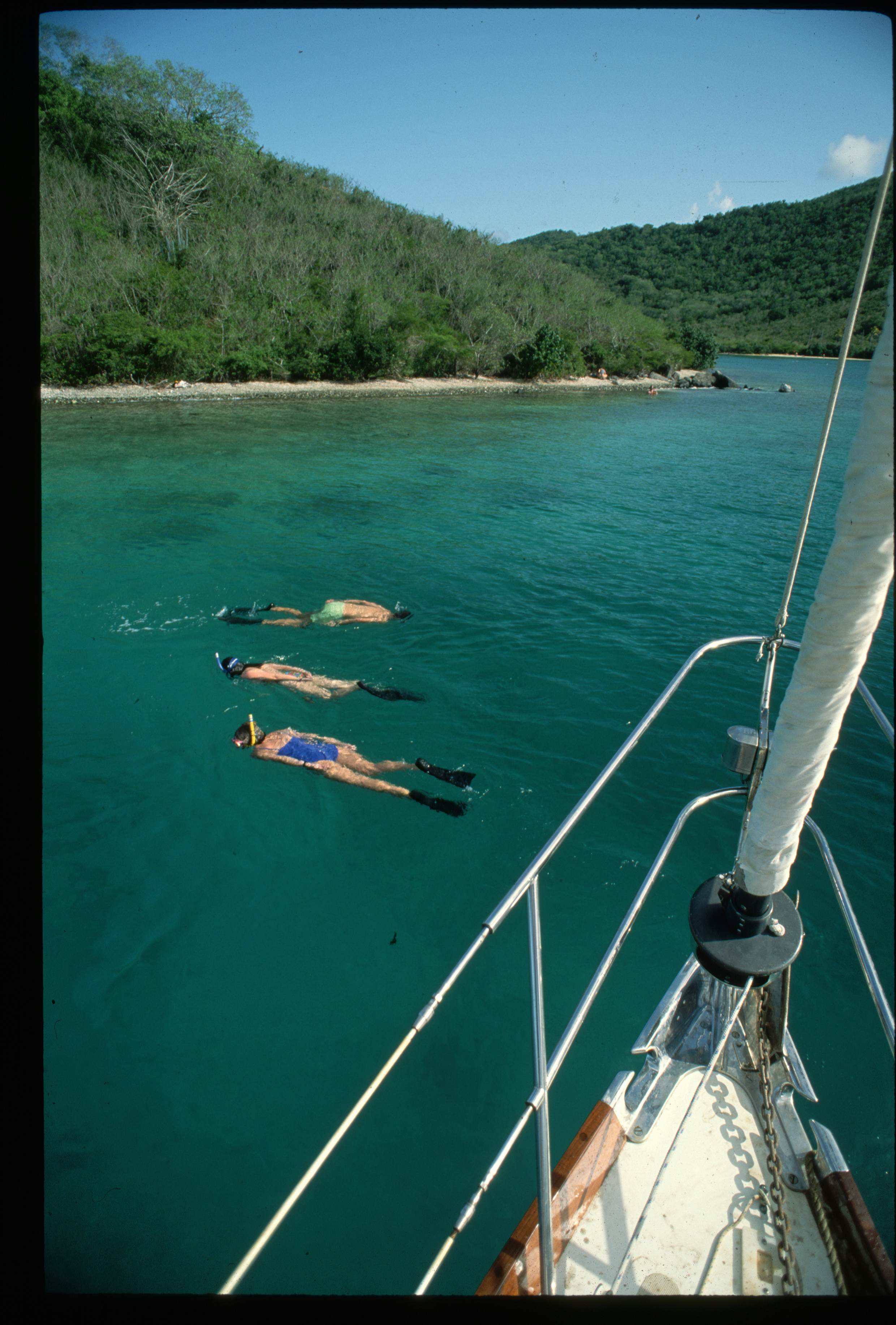 Three people snorkel in blue-green water beyond a sailboat's prow on a sunny day.