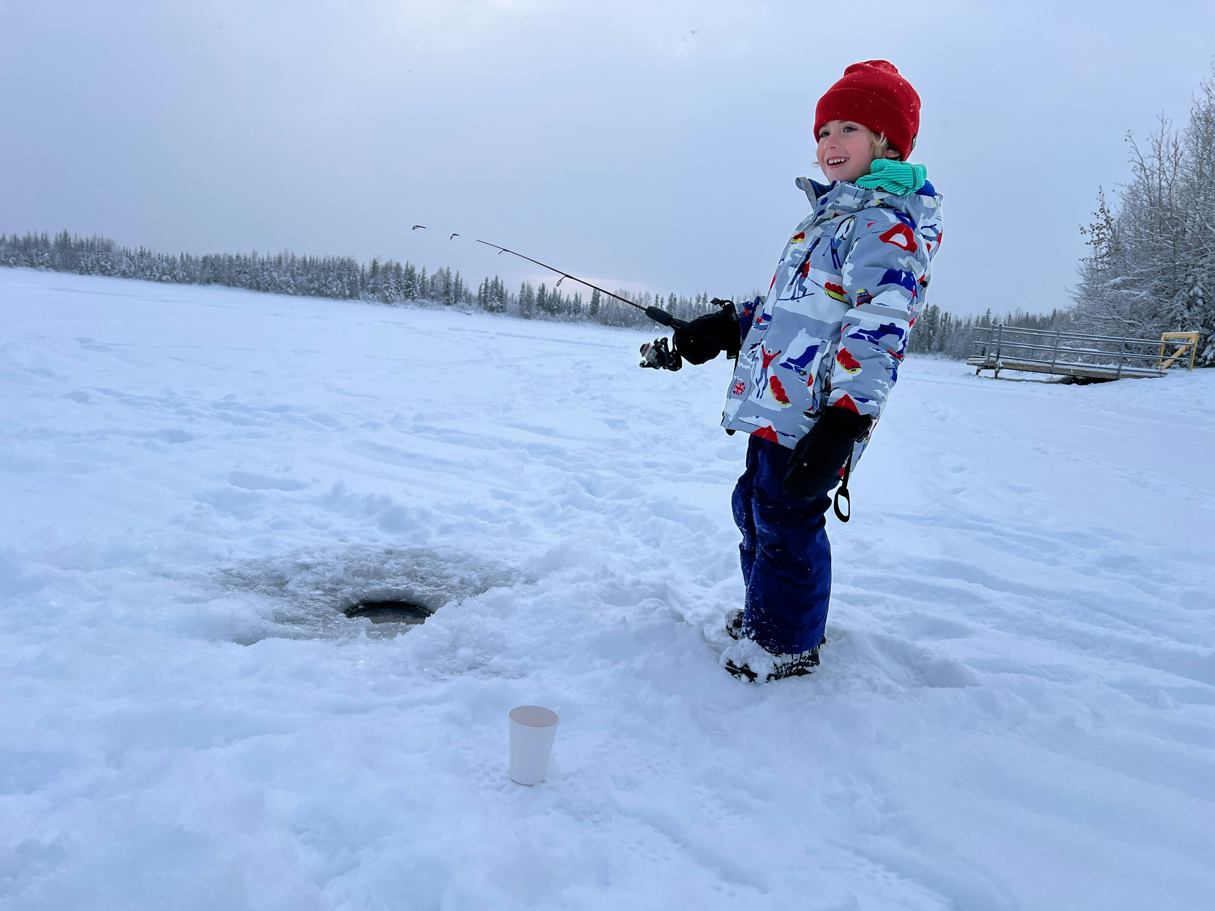 A boy wearing a red had, blue pants and a gray patterned jacket holds a fishing pole over a hole in snow-covered ice in Alaska.