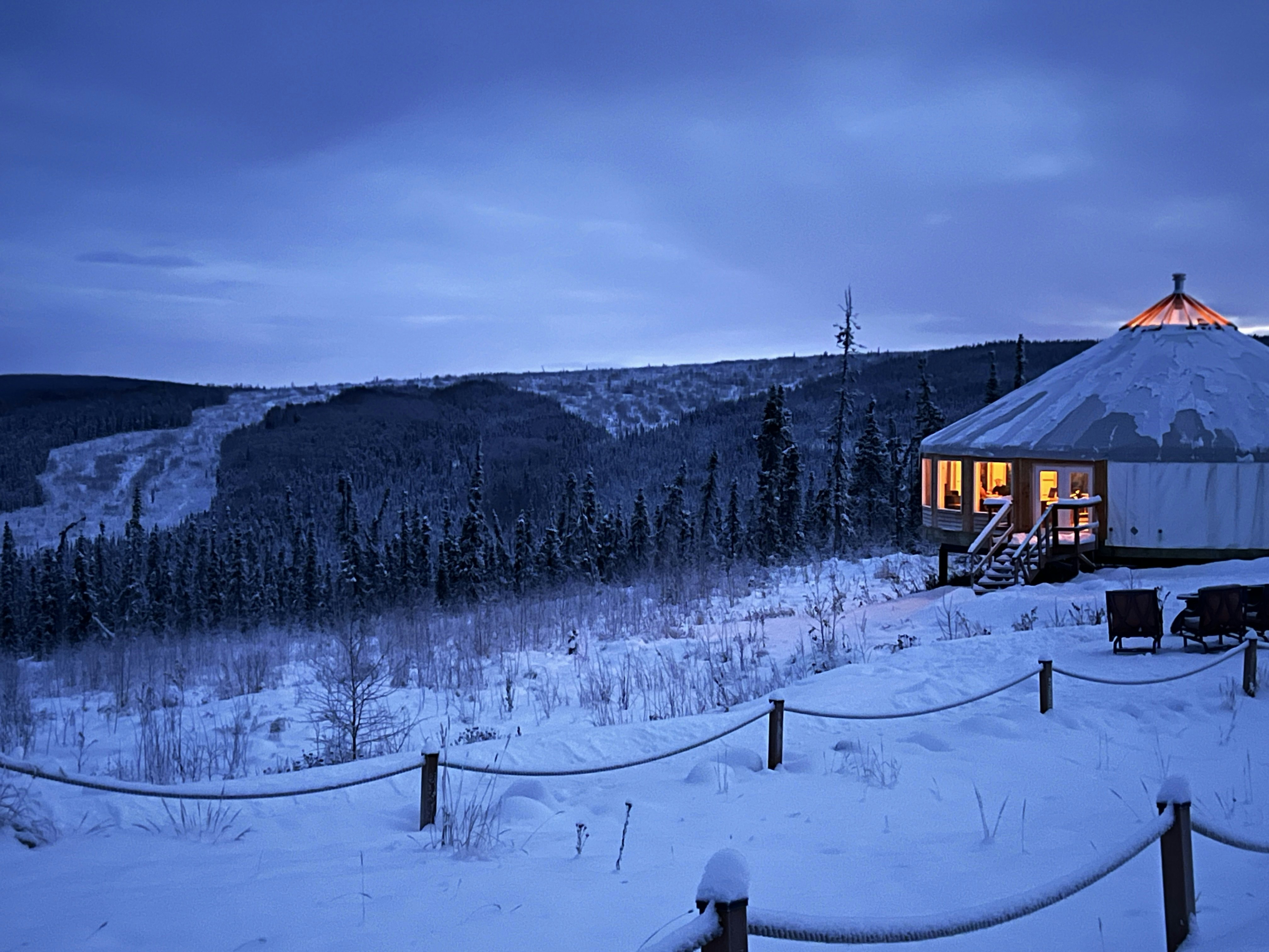 A large yurt lit from within in a snowy landscape in Alaska; two rope fences are in the foreground, and an evergreen forest is in the background.