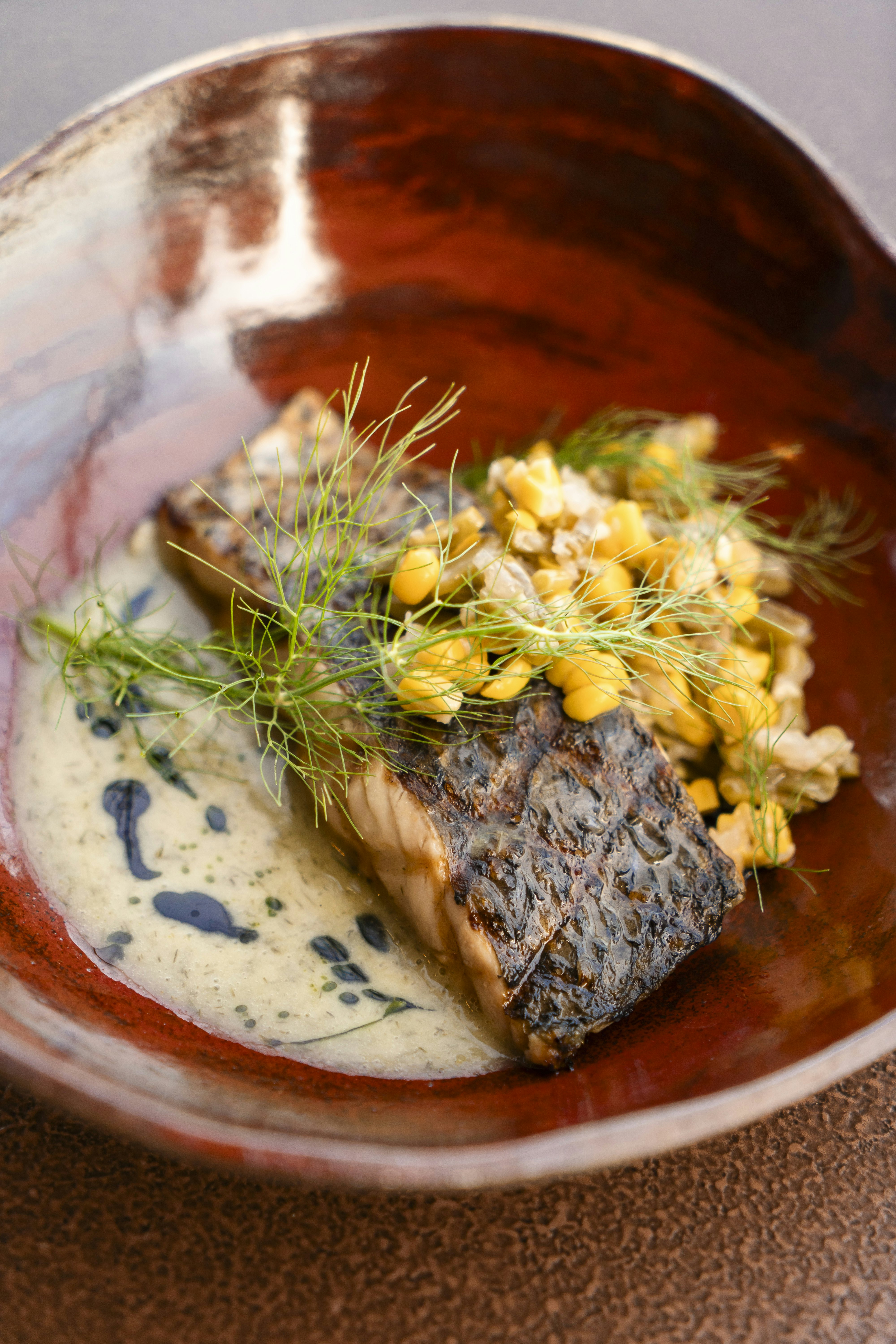 A small ceramic dish with a piece of seared fish in it topped with a white sauce and sweetcorn.