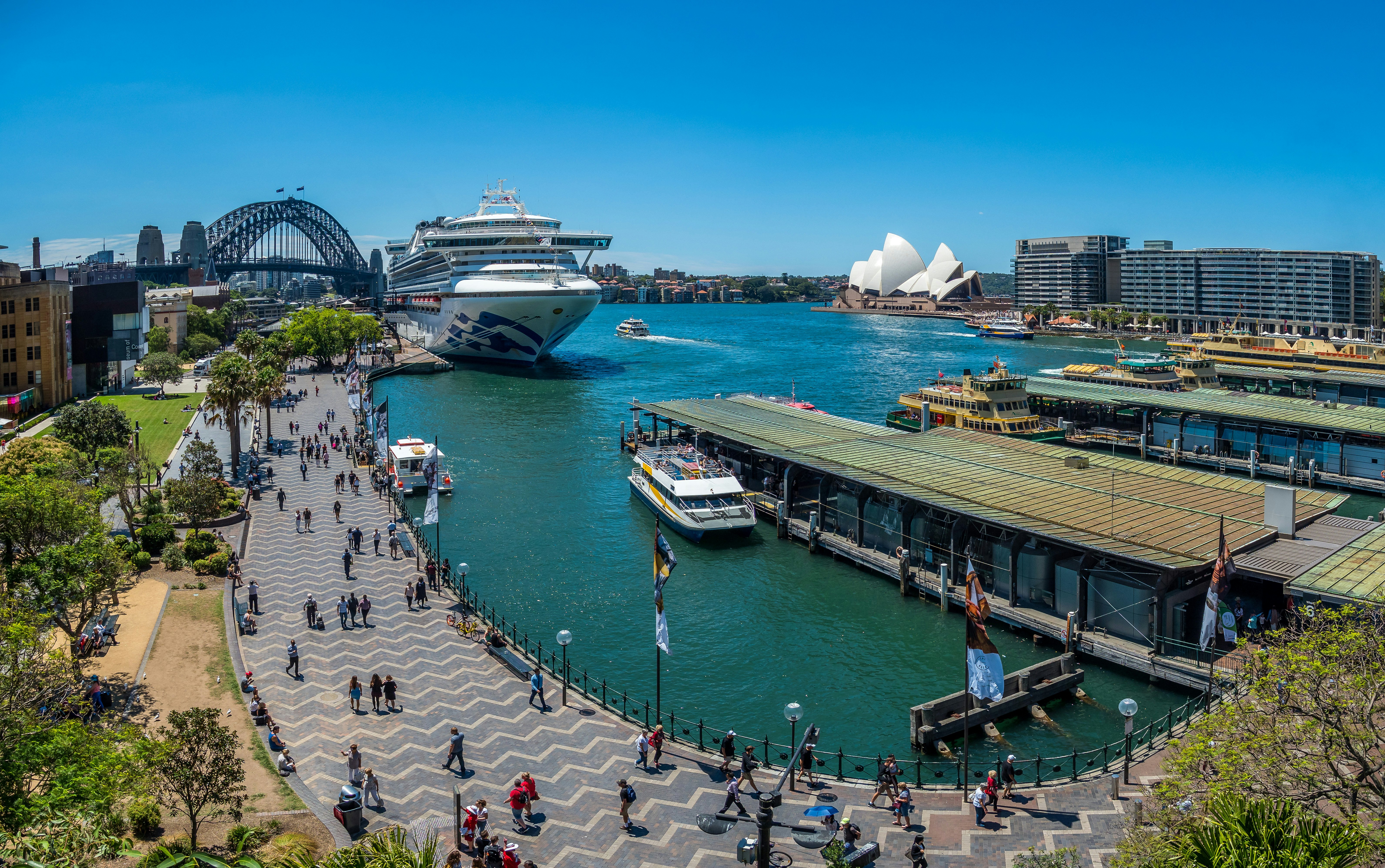 A busy harborfront. A huge cruise ship has docked near a large bridge that looks like a coathanger; a white building with a sail-shaped roof stands on the edge of the quay.