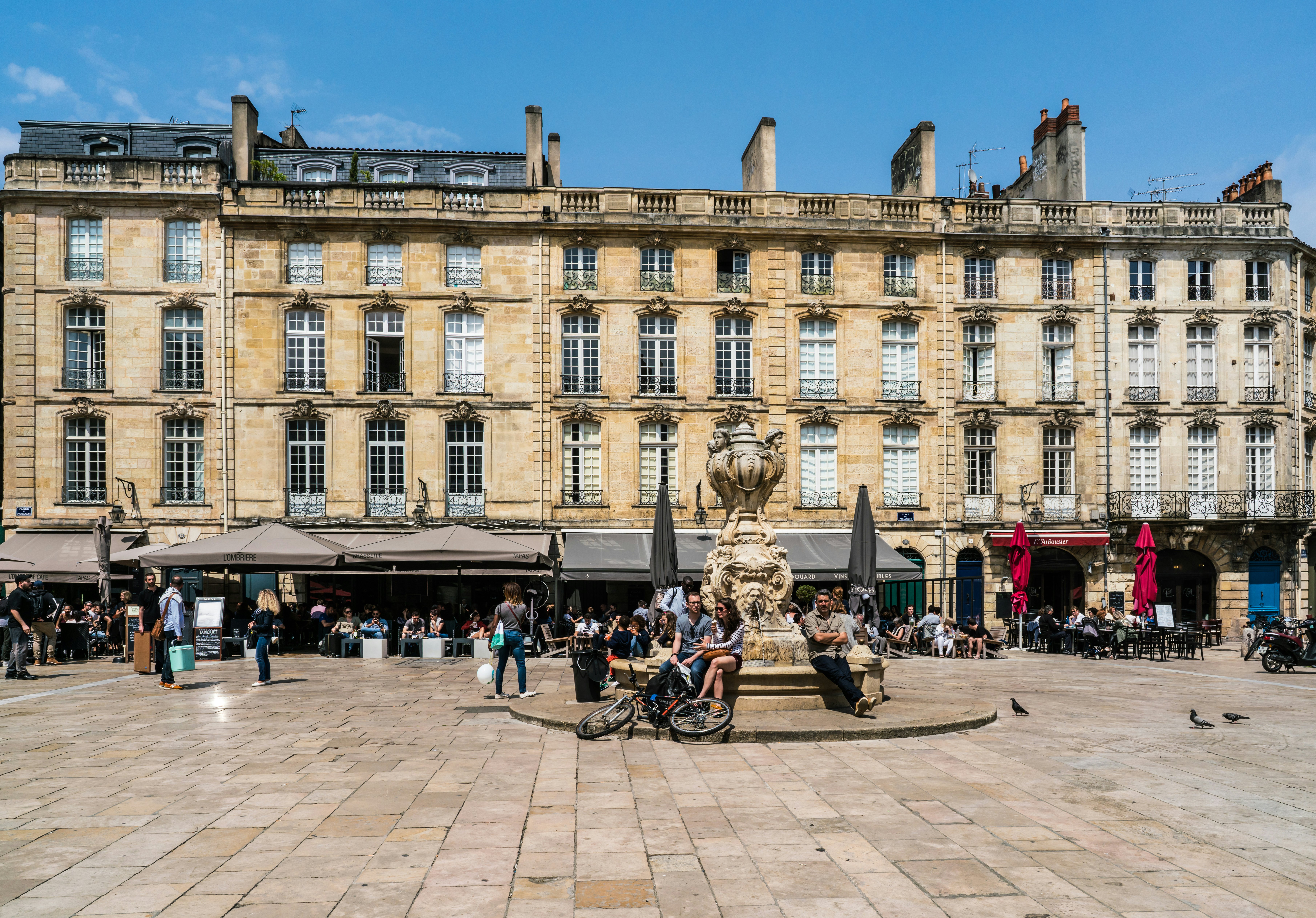 People sit around a fountain and at restaurant terraces in a city with grand architecture.