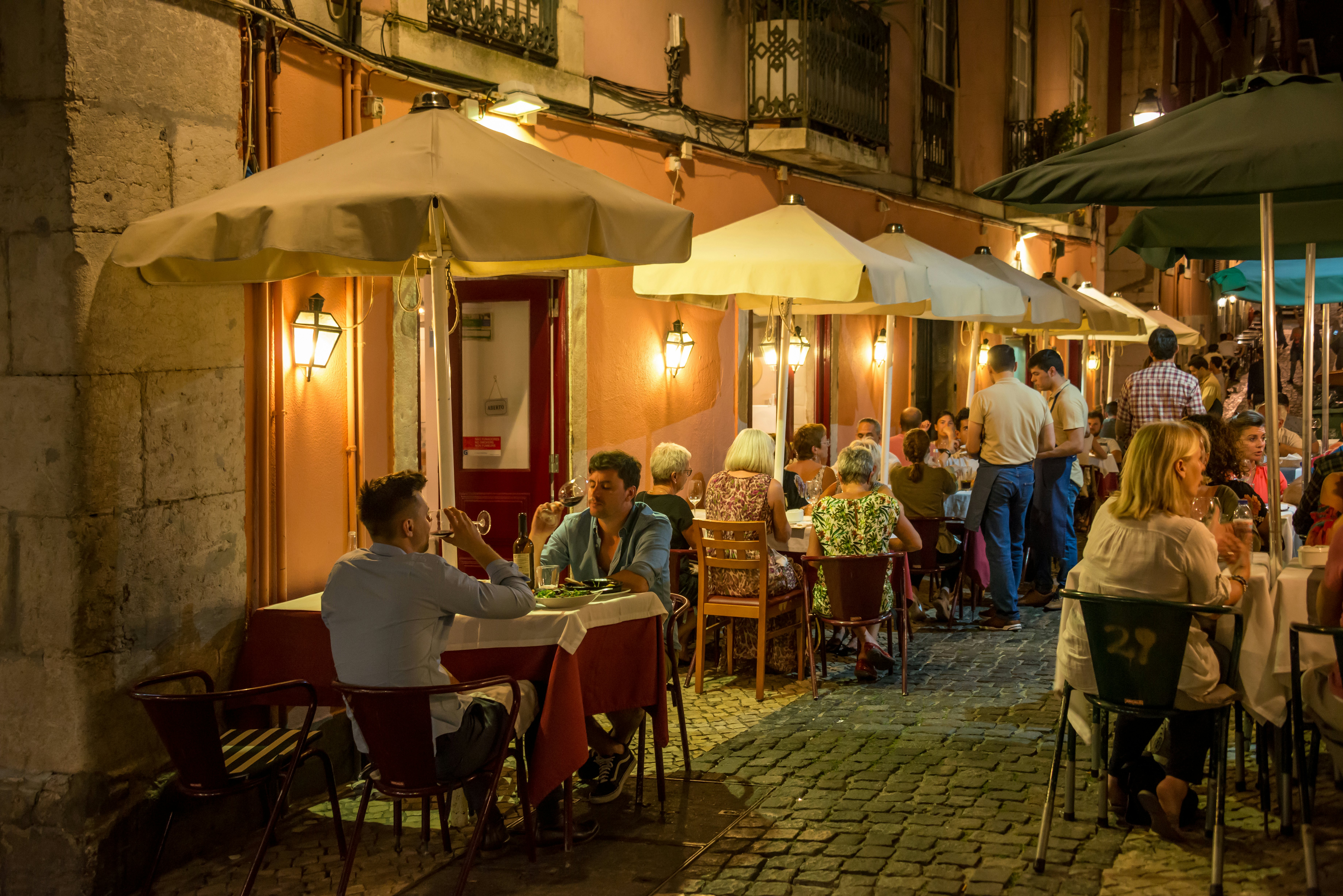 People sit at tables at an outdoor cafe along a cobblestone alley at night.
