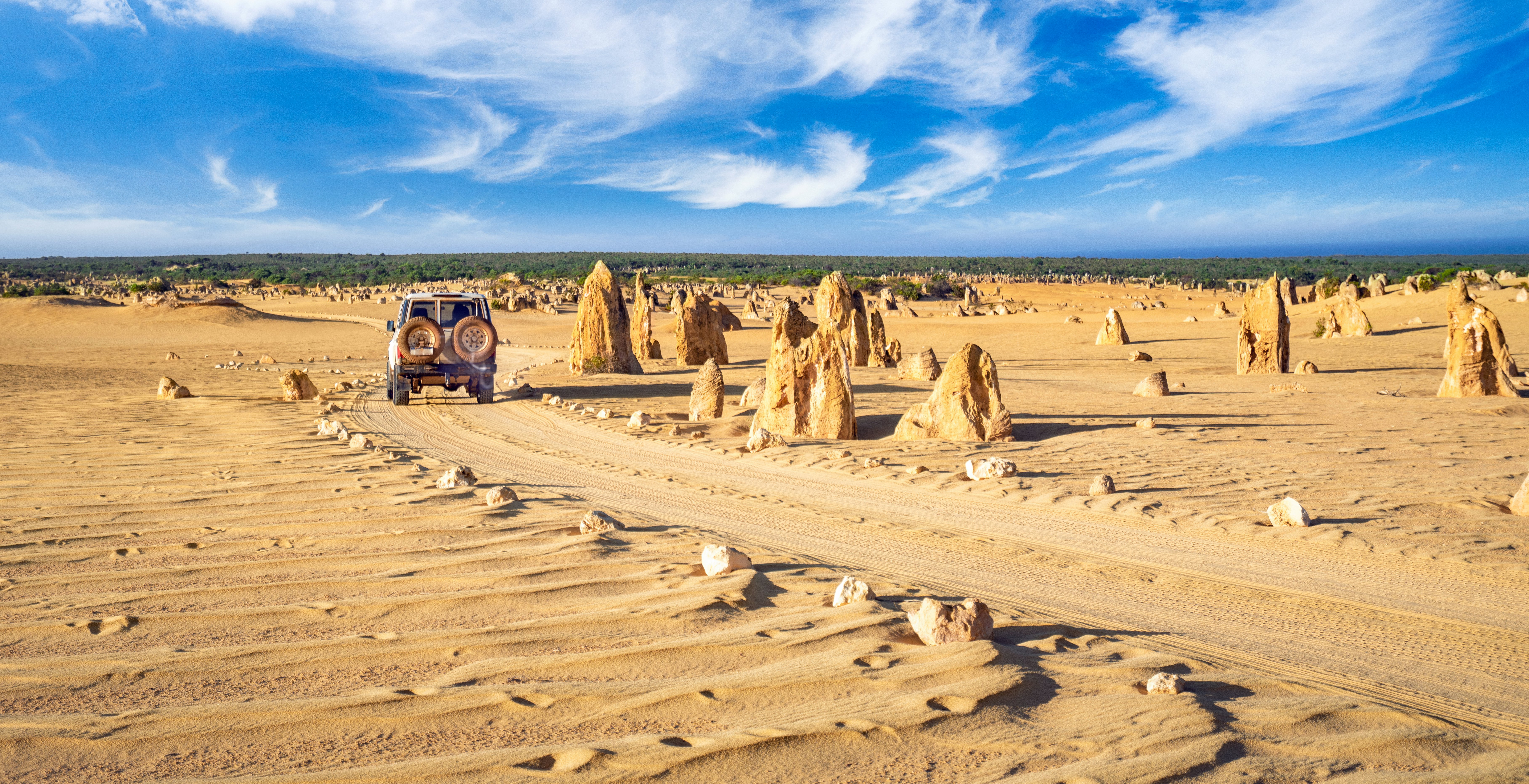 A four-wheel drive vehicle follows a sandy track through pointed rock formations that jut straight out from the sand.