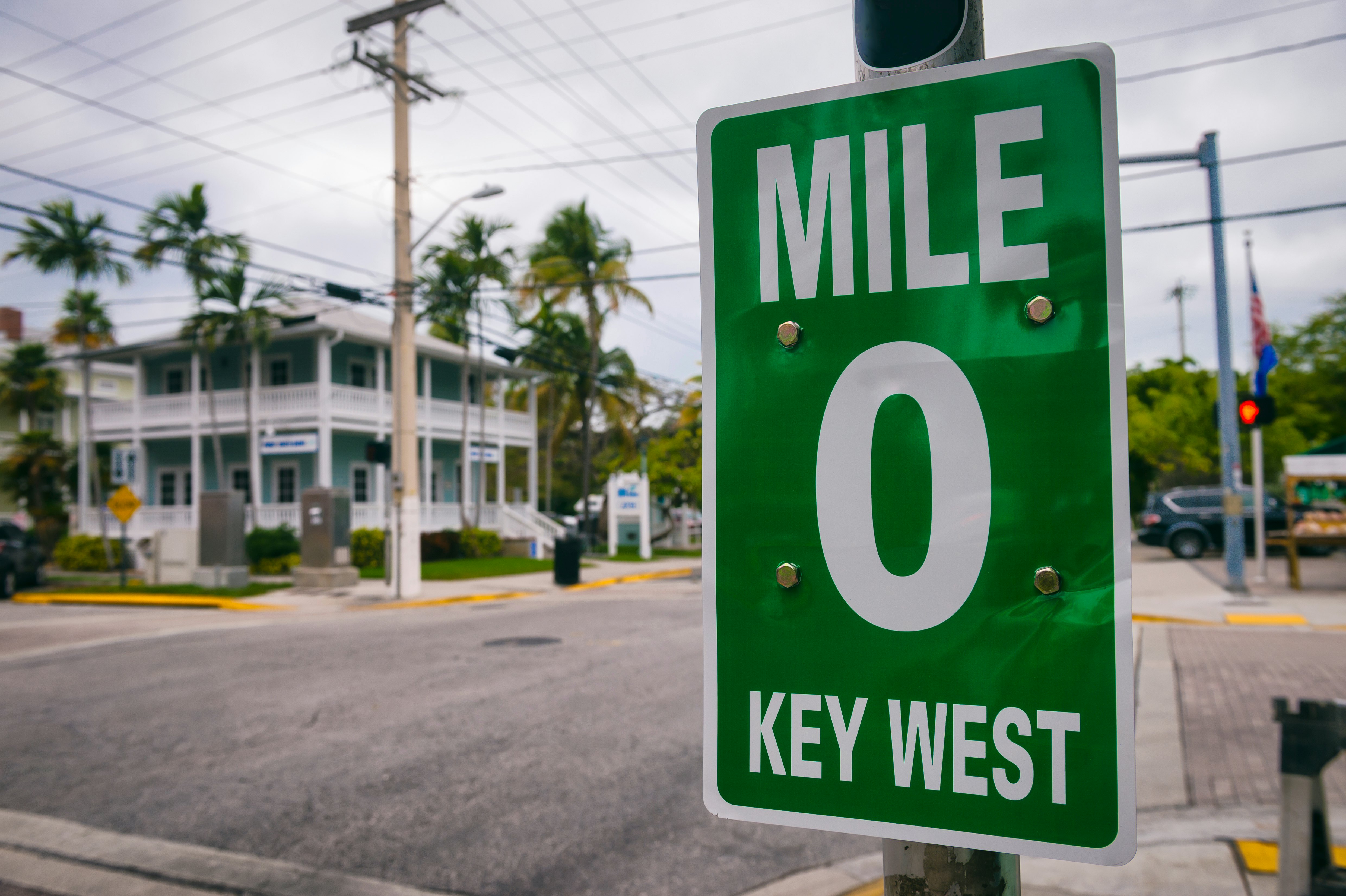 A road sign denotes “mile 0” along a city street. A building with white-painted porches is seen across the street.