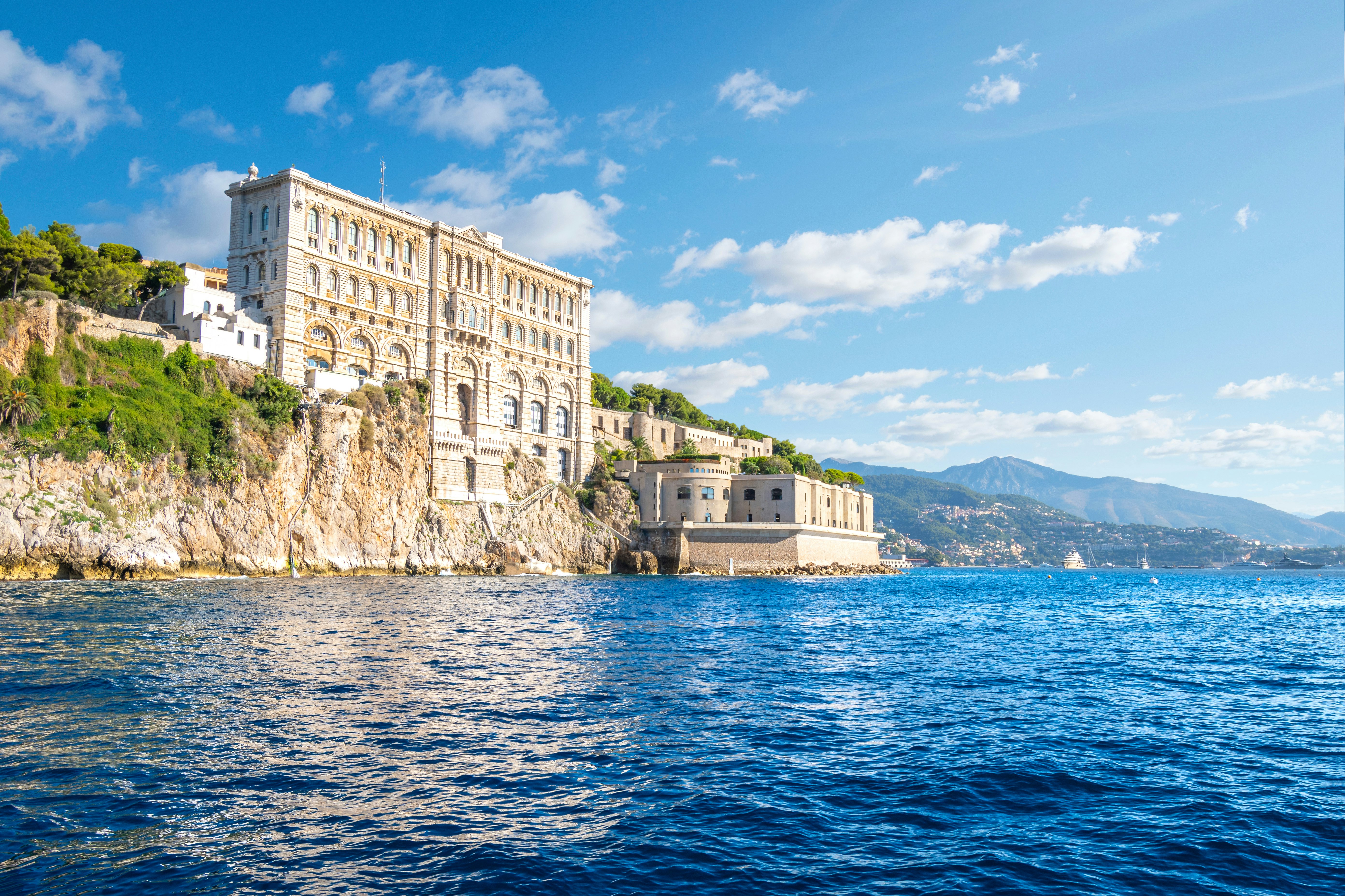 Looking from the ocean toward a tall stone building with mountains in the right distance on a sunny day.