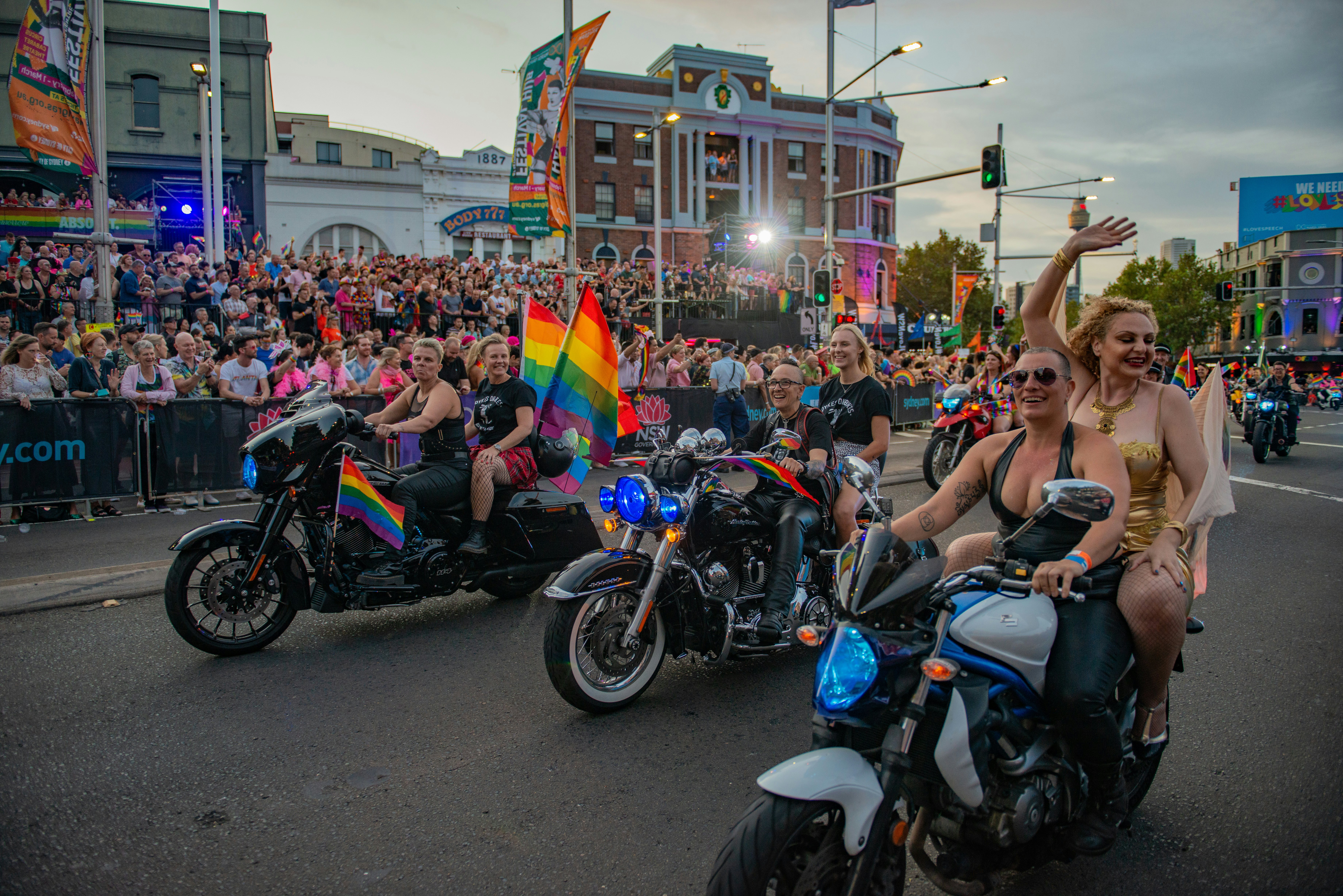 People on motorcycles with rainbow flags on the back wave at spectators on a parade route
