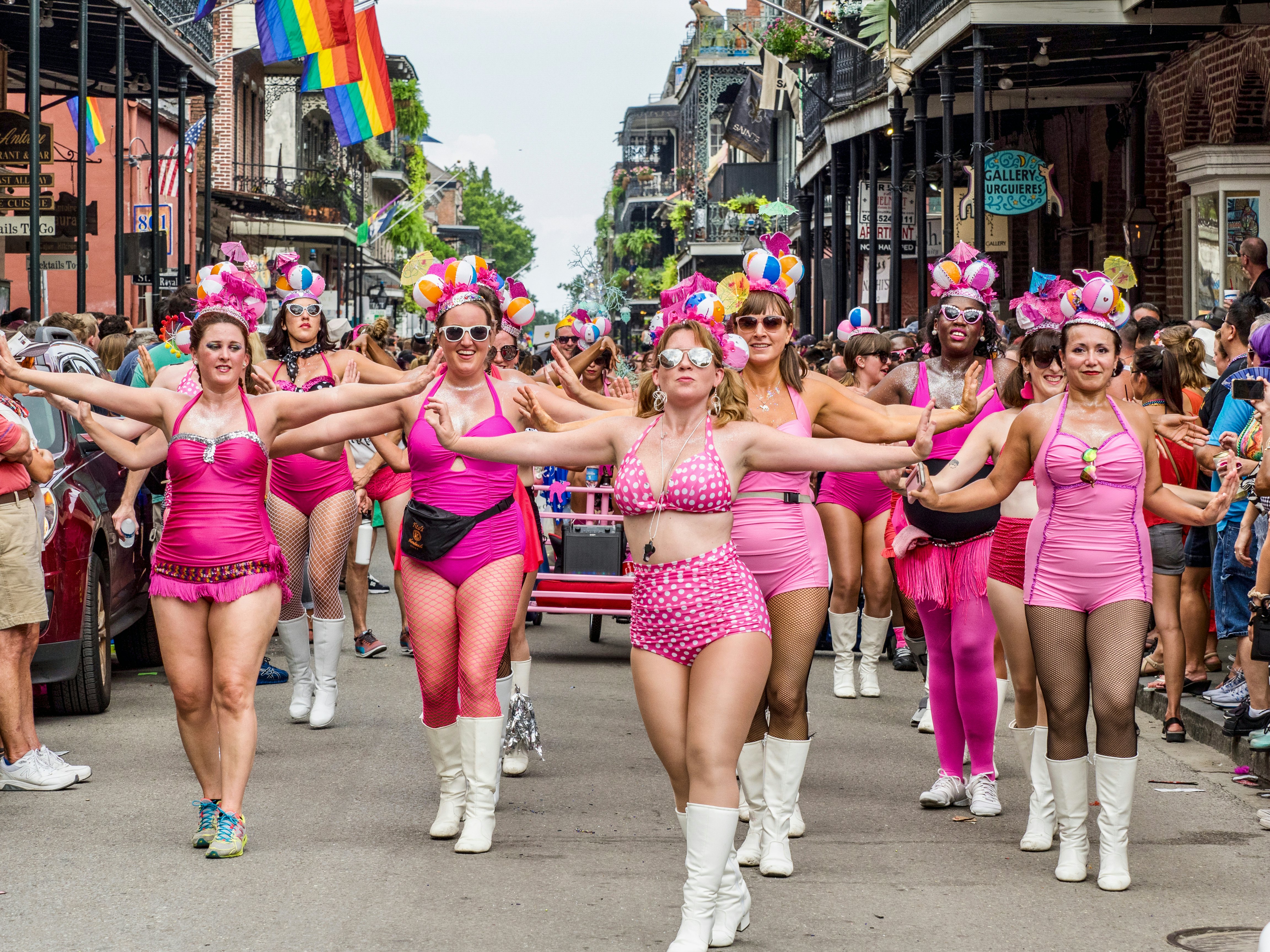 Women in pink outfits march in formation on a street in a historic district in a city during an LGBTQ+ celebration.