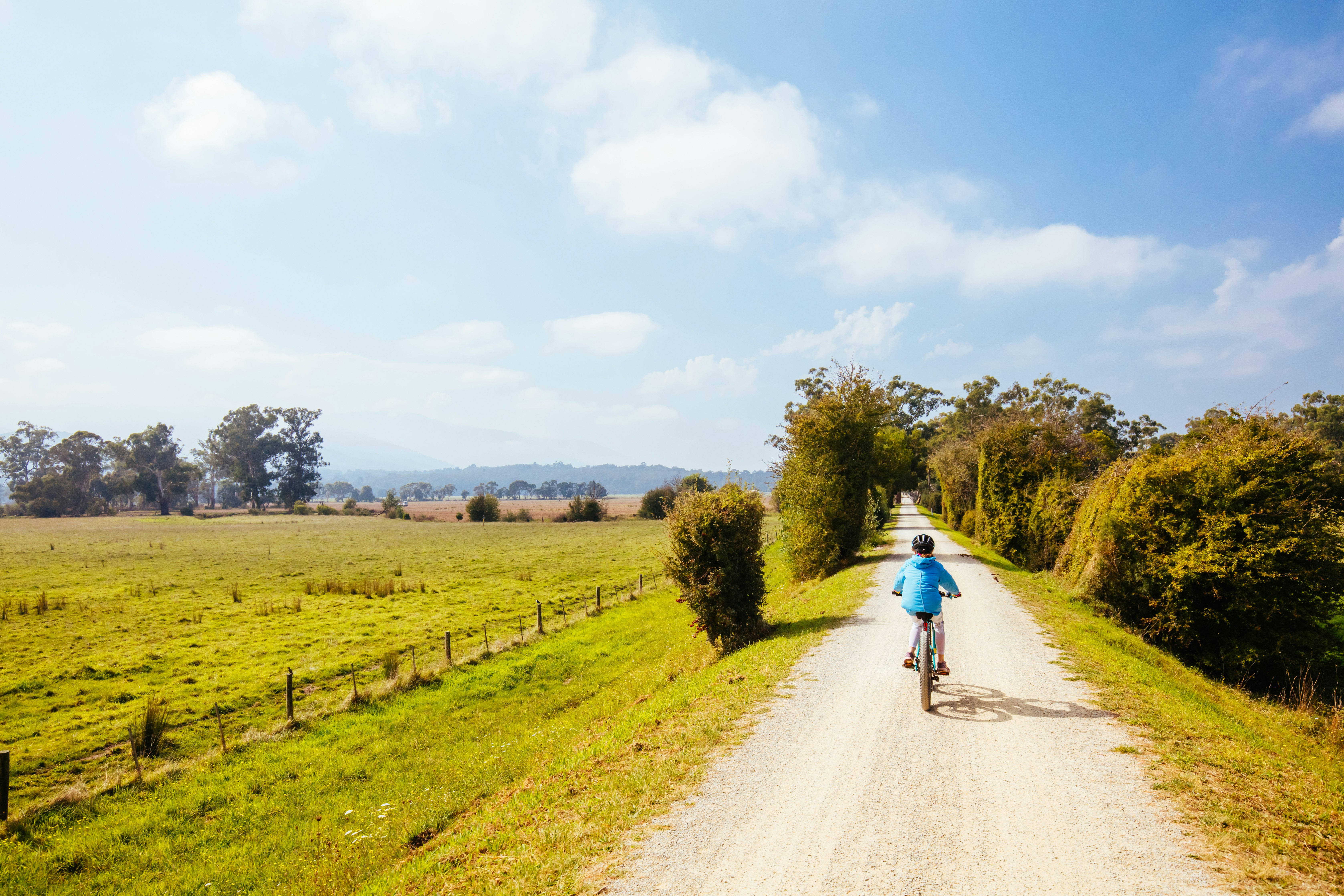 A child cycles on a narrow, straight walking path runs through green fields between Lilydale and Warburton in Australia.