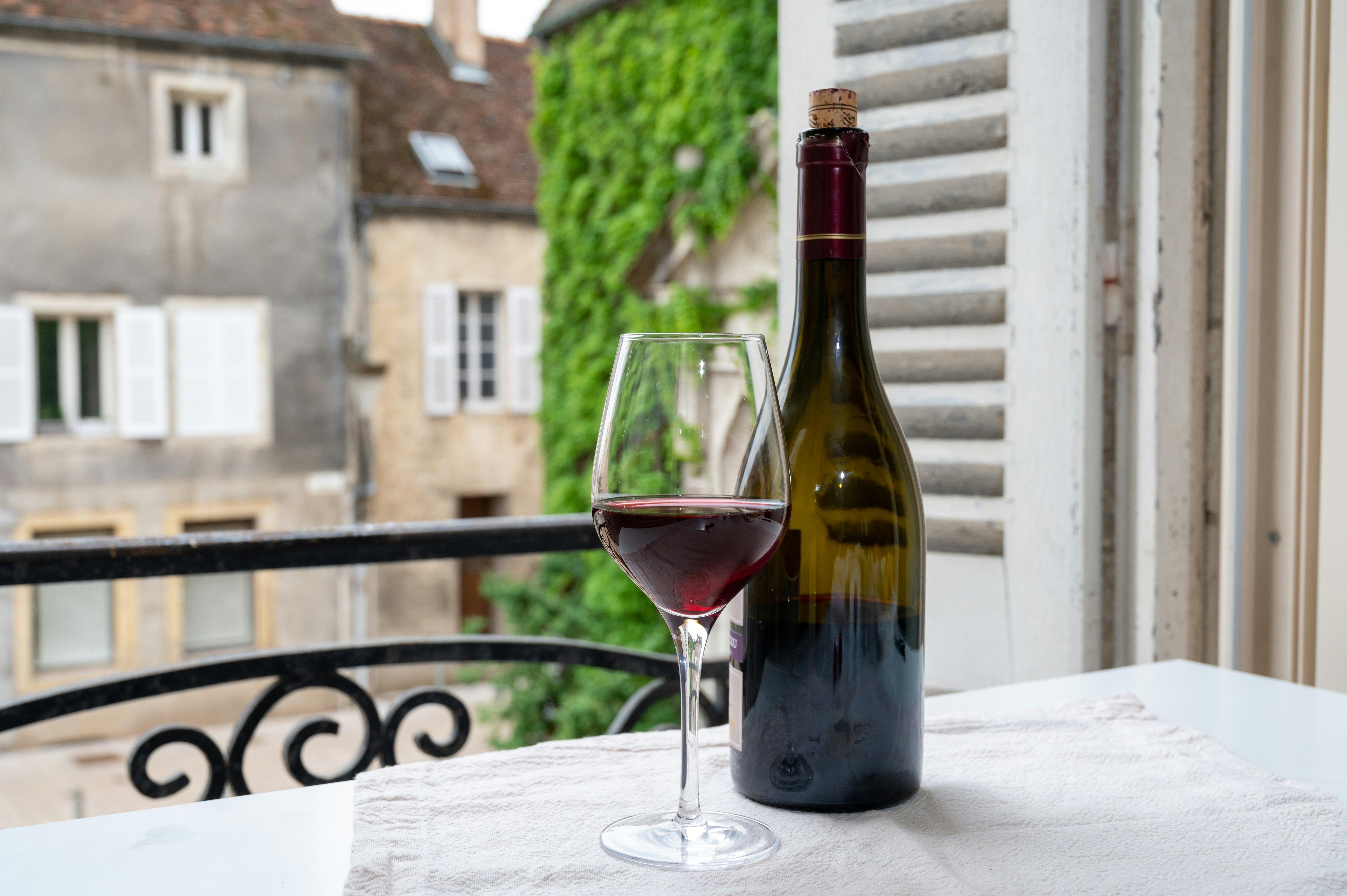 A bottle of red wine and a glass stand on a table near a balcony window in a small village.