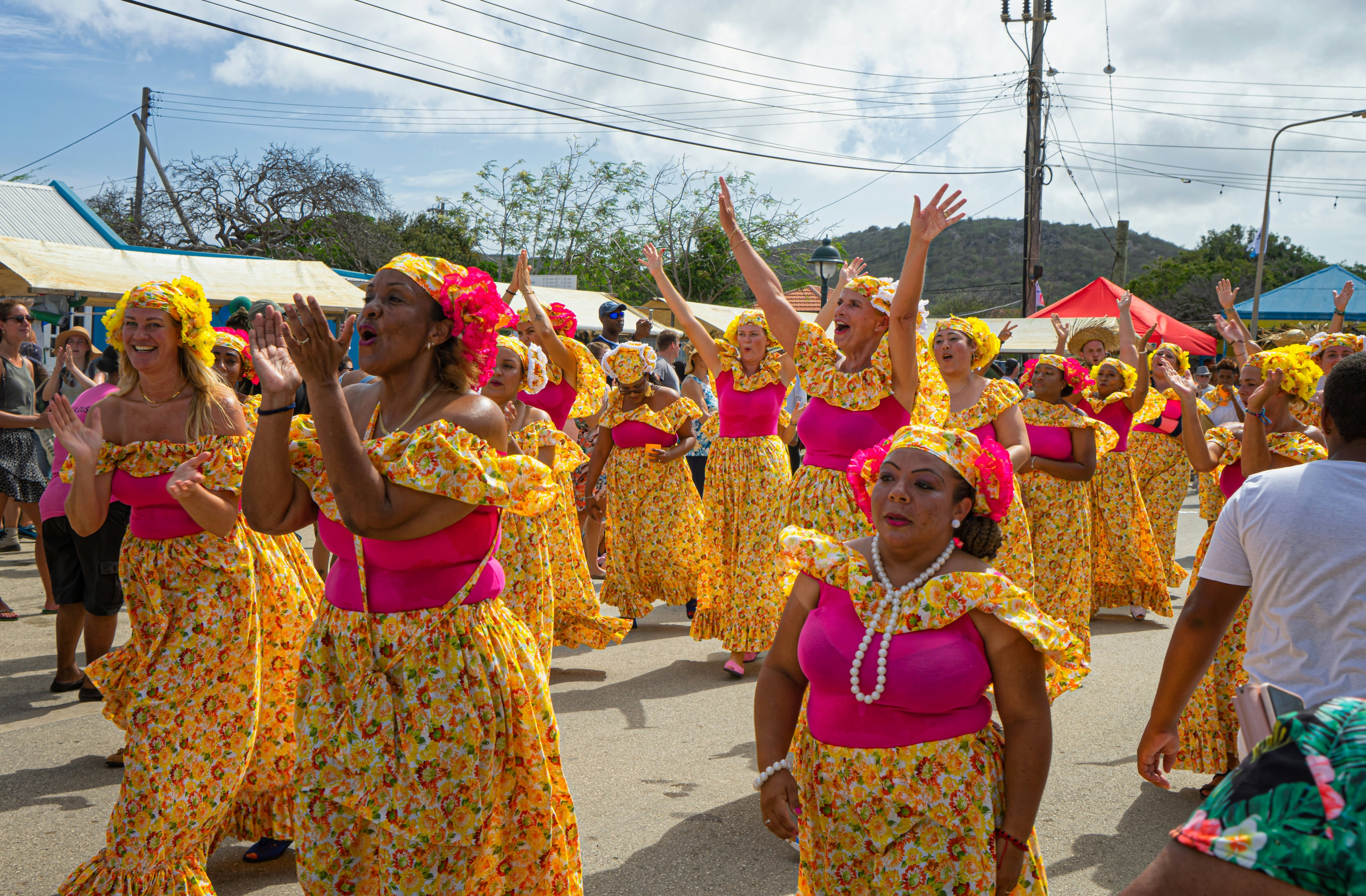 Women in matching outfits of hot pink tops and patterned yellow skirts and scarves march in a parade in a small town.